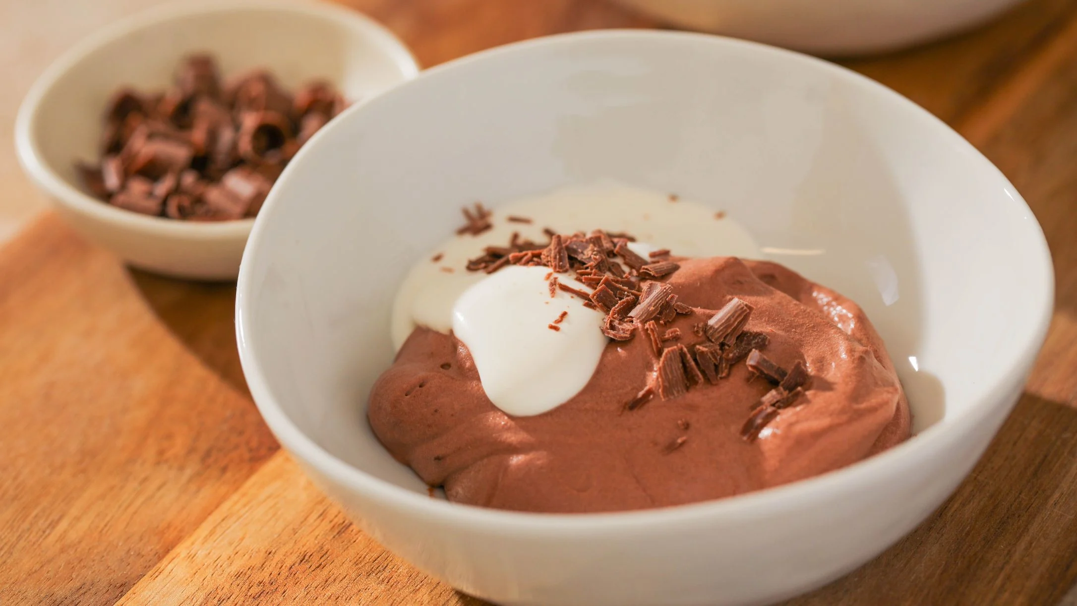 A white bowl of chocolate mousse topped with cream and chocolate shavings, with a small bowl of shavings nearby.