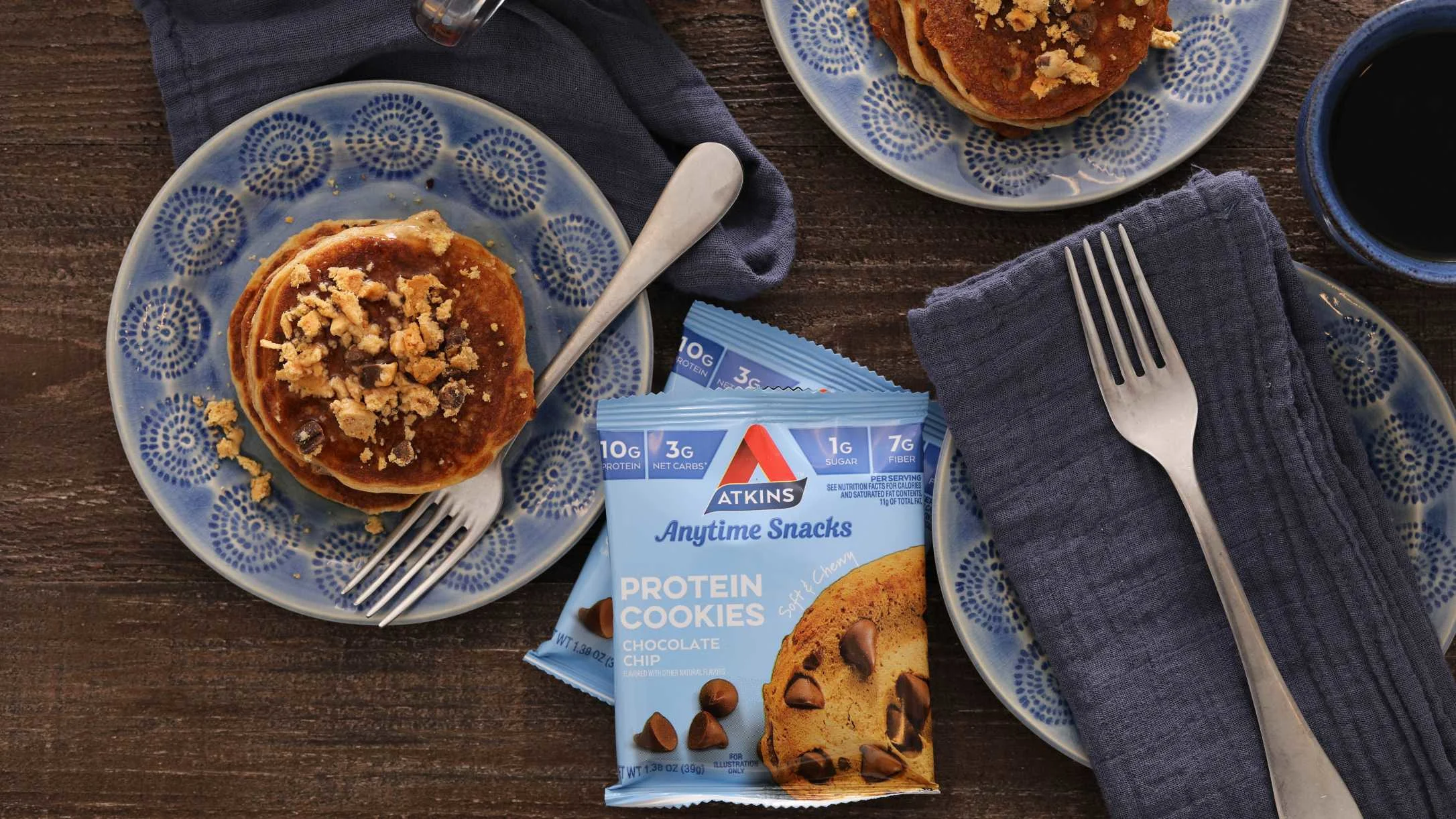 Atkins Anytime Snacks Protein Cookies package beside pancakes on blue plates with forks and coffee on a wooden table.