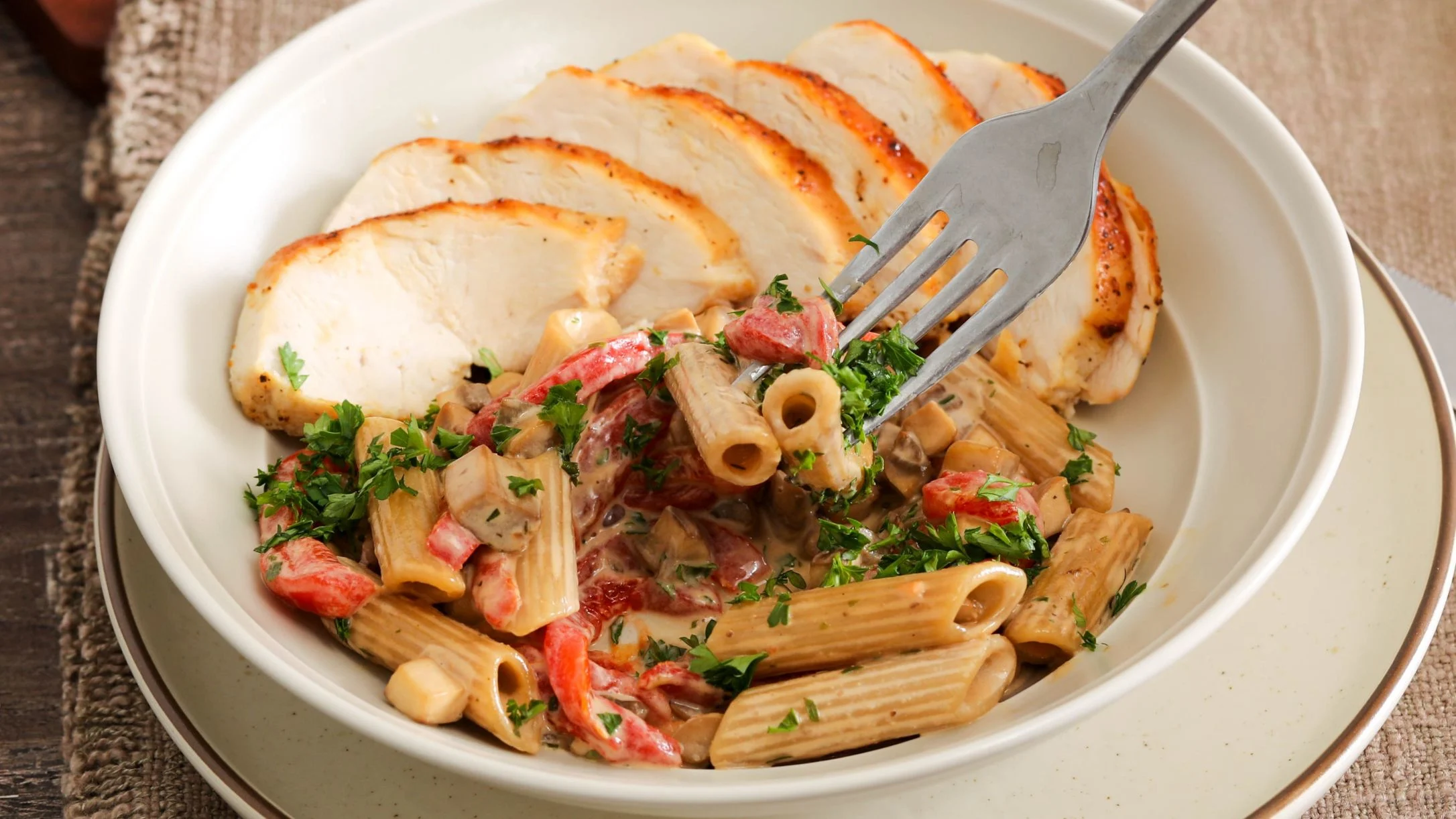 A white bowl with sliced grilled chicken and penne pasta in creamy sauce with roasted peppers, mushrooms, and fresh parsley, with a fork.