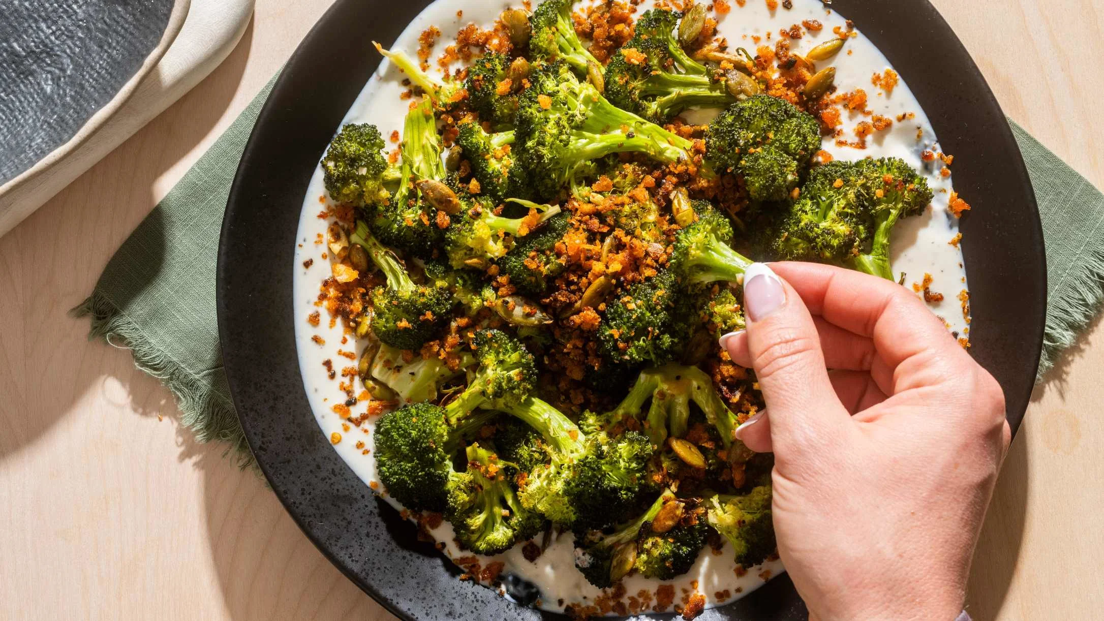 Roasted broccoli florets on a white plate with spiced breadcrumb topping, hand reaching to serve.