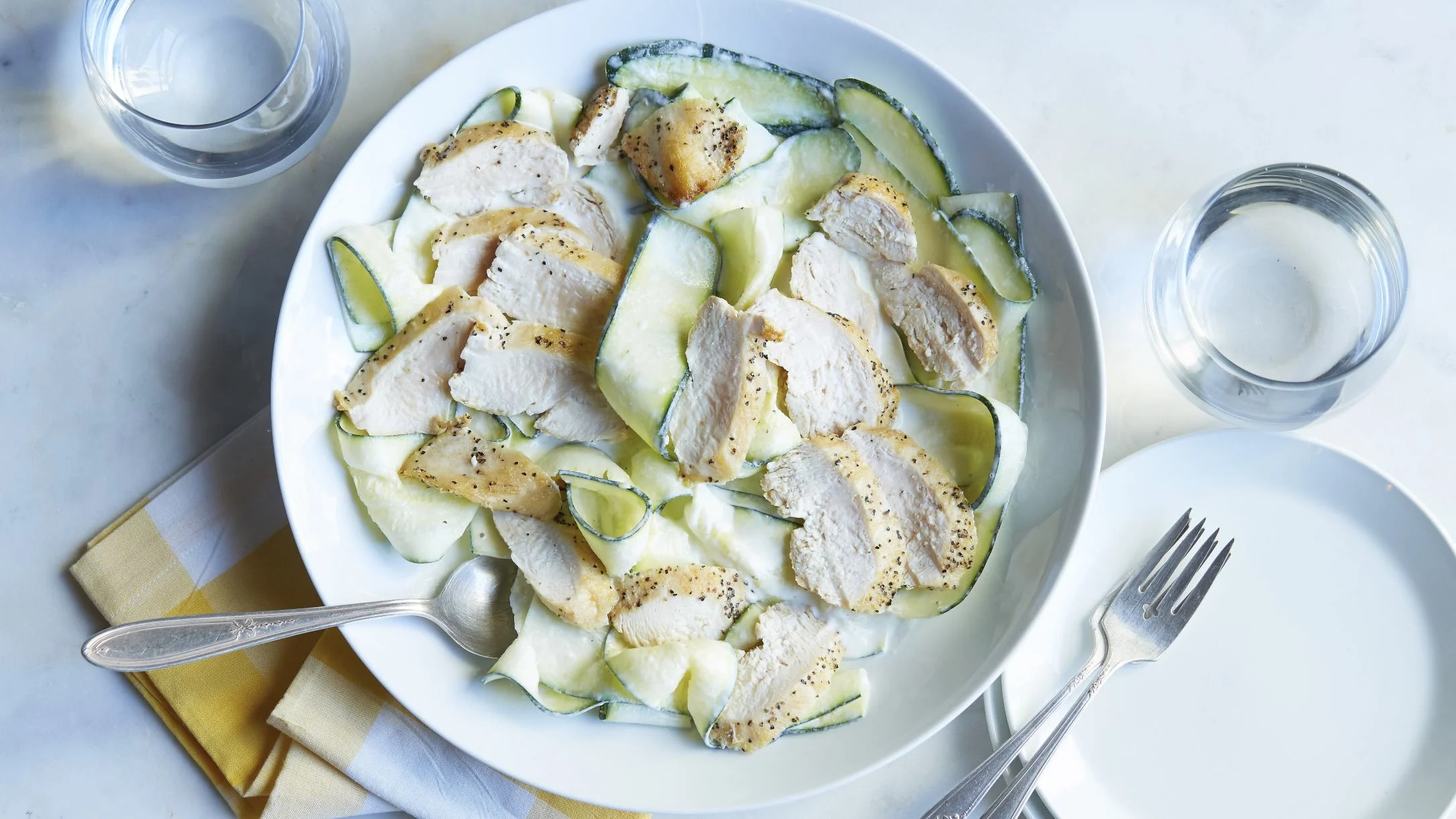 A white bowl with sliced seasoned chicken over zucchini ribbons in cream sauce, with silverware, yellow napkin, and water glasses.