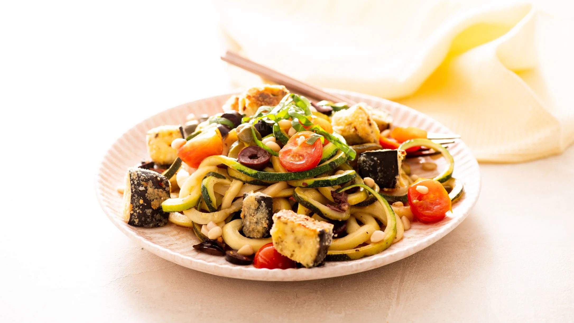 Colorful pasta dish with zucchini spirals, cherry tomatoes, olives, and eggplant cubes on a white plate with fork.