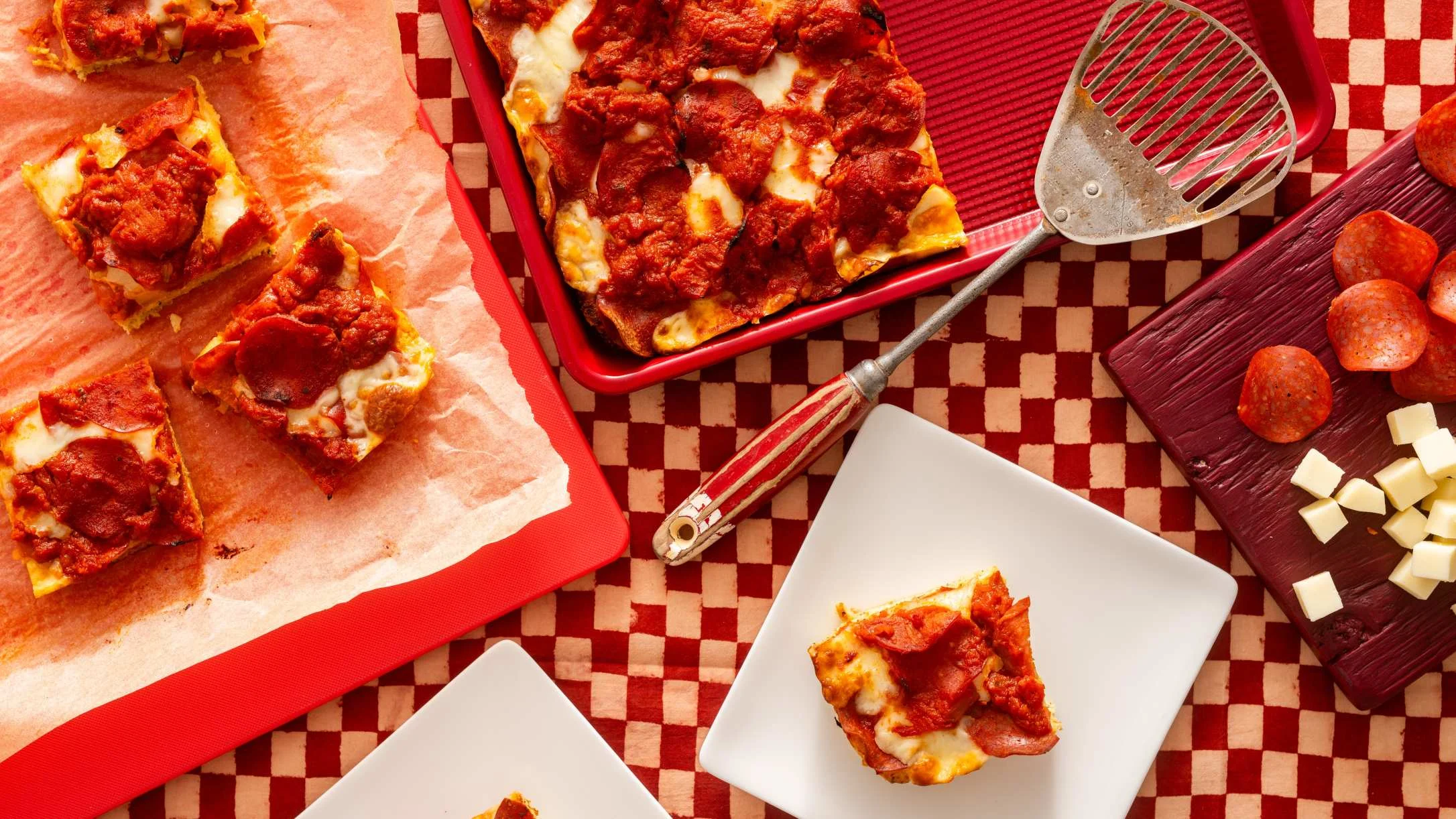 Detroit-style pepperoni pizza served on red trays and white plates with cheese cubes and pizza cutter on checkered tablecloth.