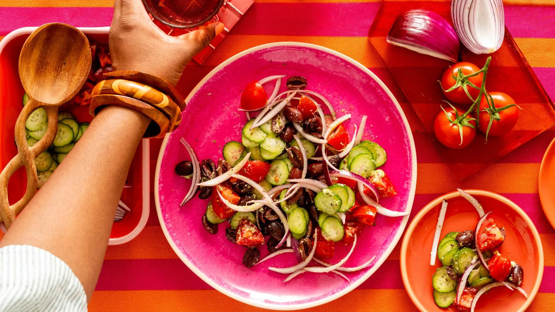 A hand holding a drink over a vibrant  salad on a pink plate, with tomatoes, cucumbers, olives, and red onion.