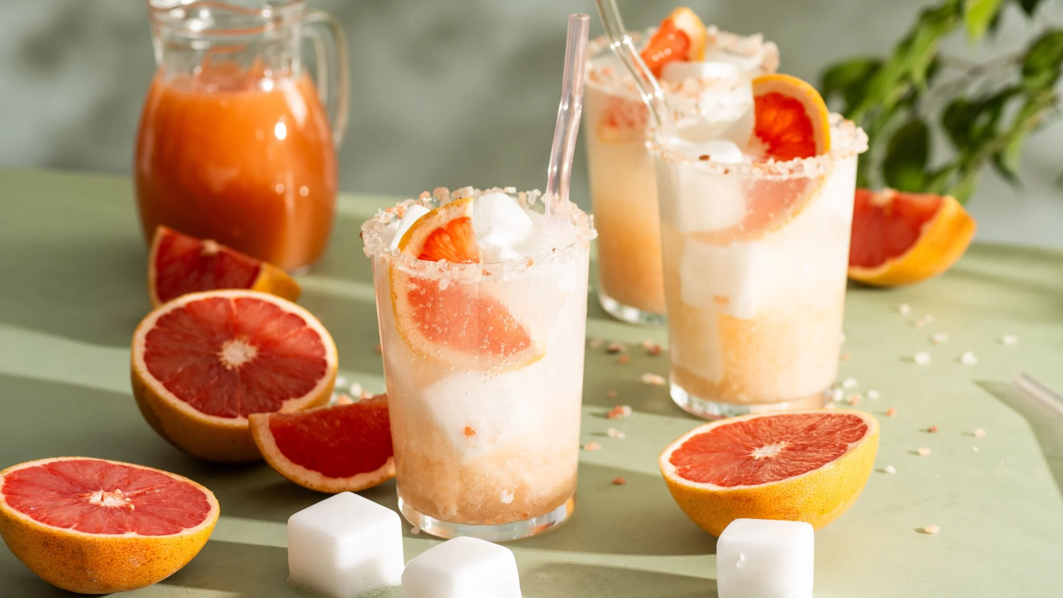 Creamy grapefruit cocktails with salt rims and fresh citrus slices, served alongside halved grapefruits and juice pitcher.