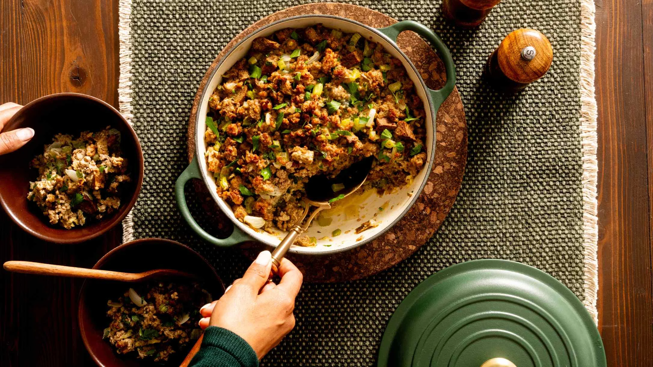A hand scoops herb-topped stuffing from a green Dutch oven into wooden bowls on a green woven placemat.
