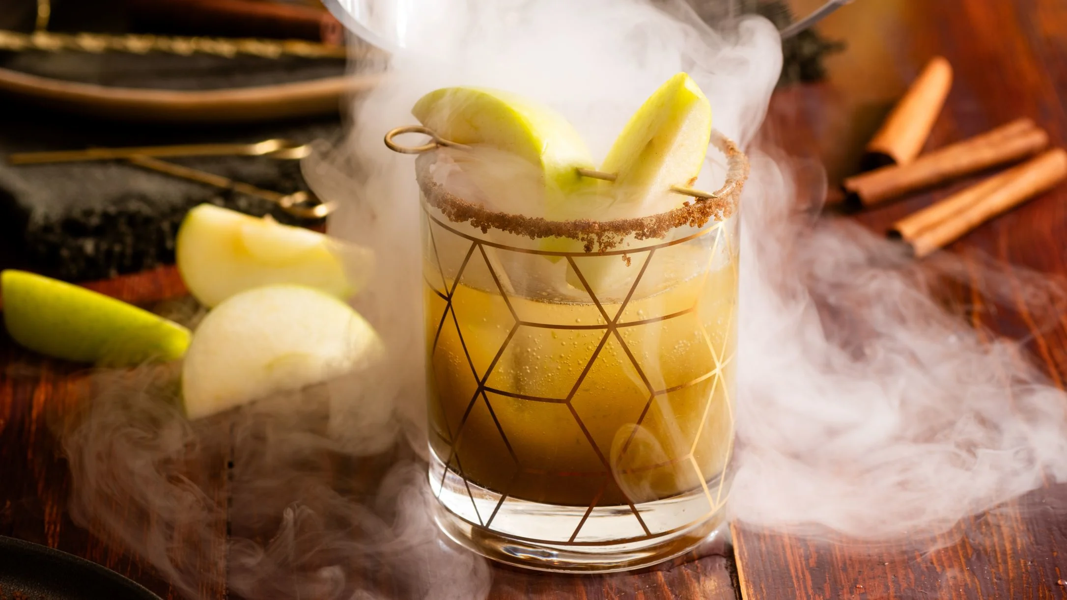 A smoky apple cocktail in a geometric glass with a cinnamon rim, garnished with green apple slices, surrounded by cinnamon stick smoke.