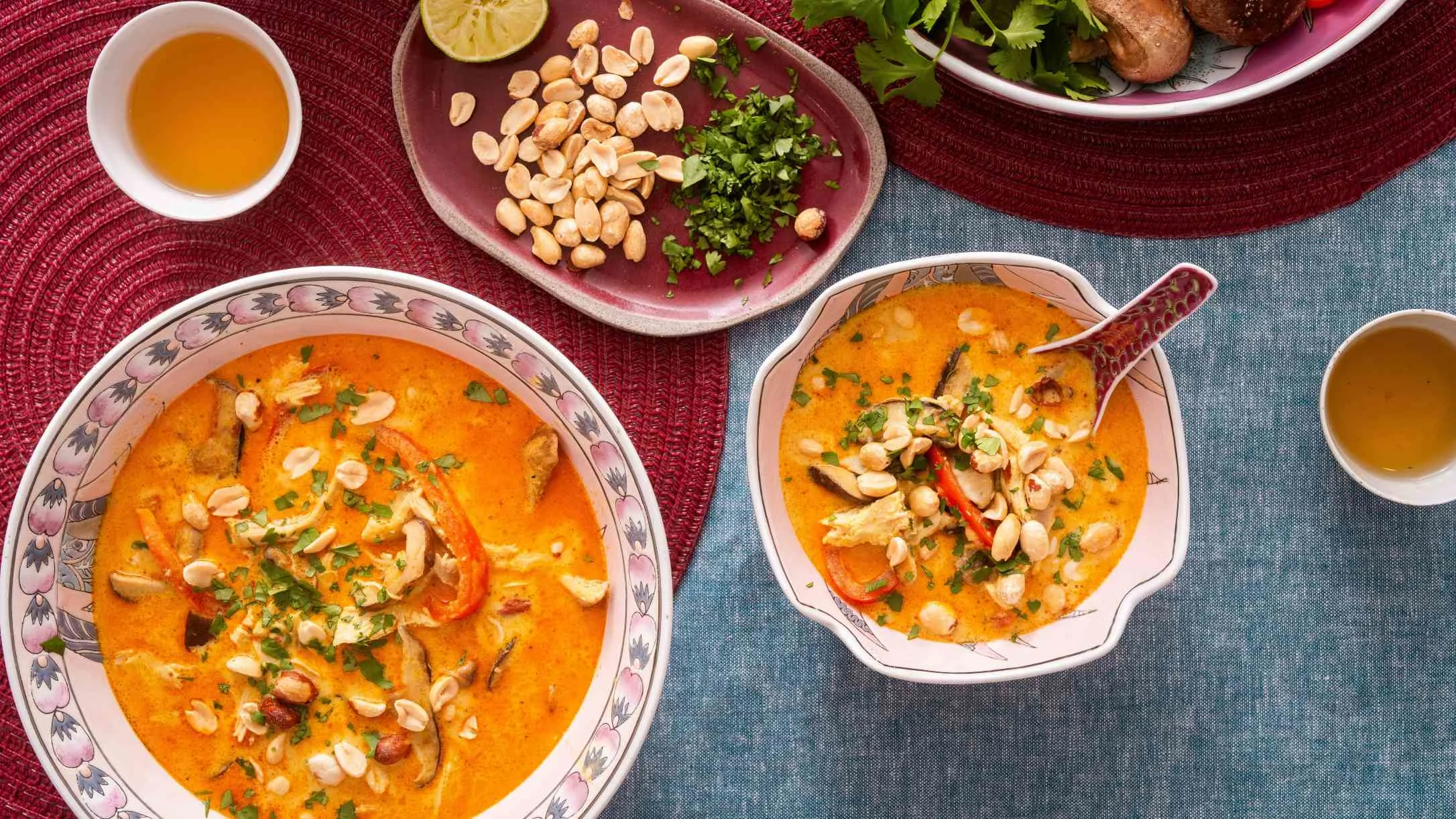 Orange curry dishes topped with peanuts and herbs, served with lime, cilantro, and tea on red placemats.