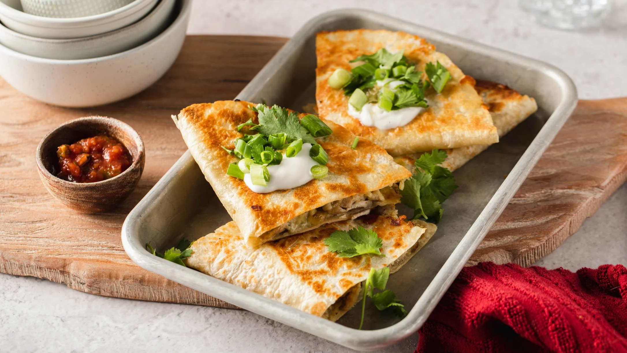 Golden quesadillas topped with sour cream and green onions on a metal tray, served with salsa in a small bowl.
