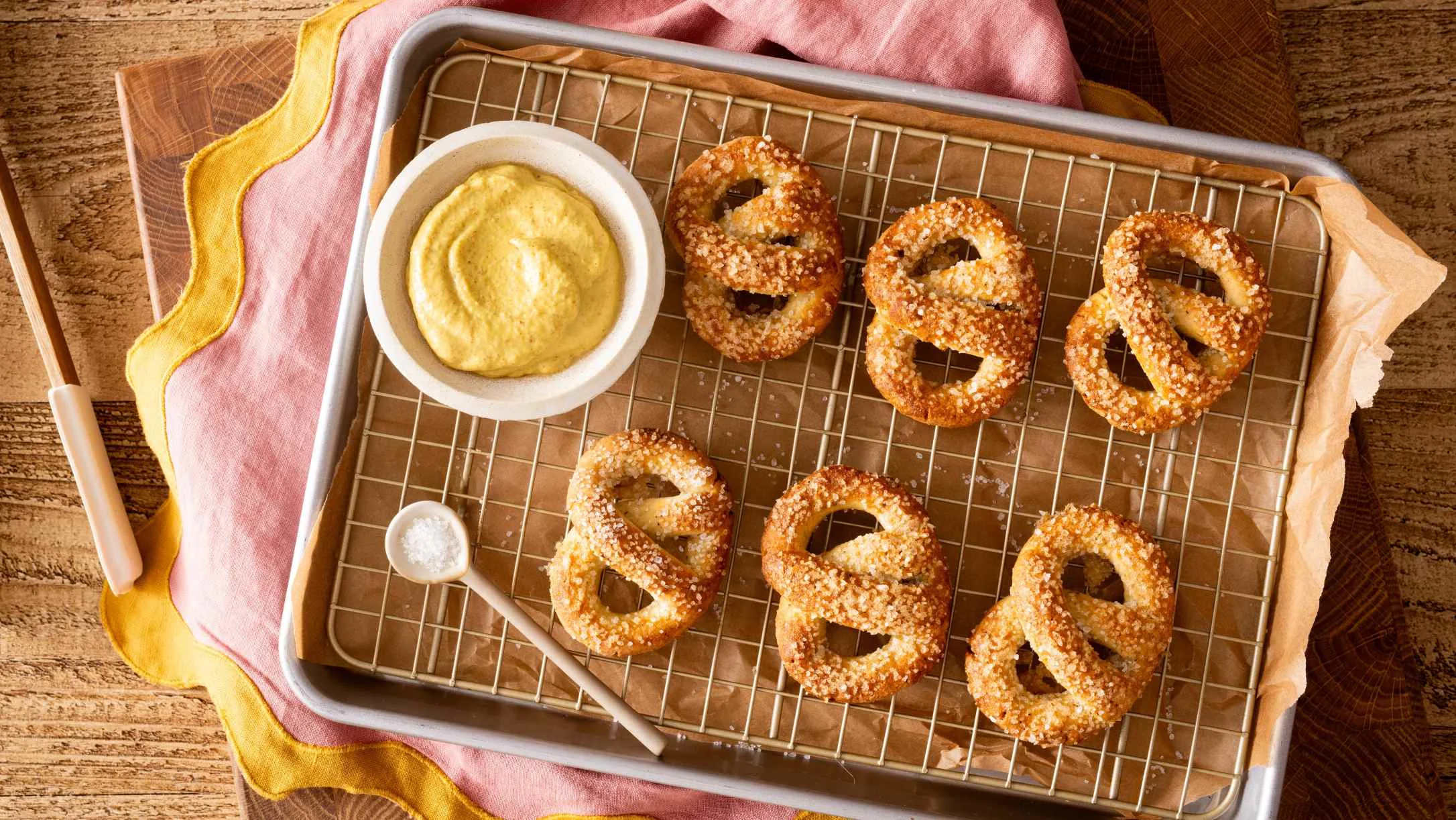 Six golden-brown salted pretzels on a wire rack beside a bowl of yellow mustard dipping sauce.