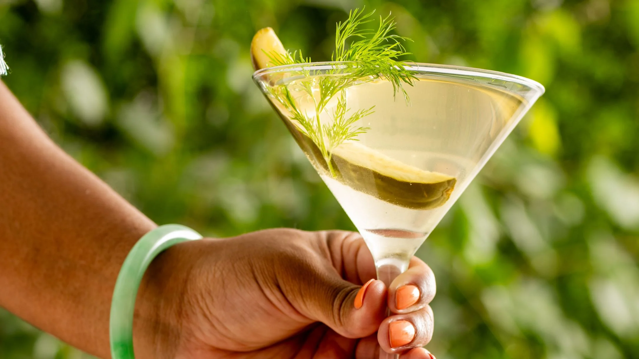 A hand with orange nails holds a martini glass garnished with dill and a pickle slice against a green leafy background.