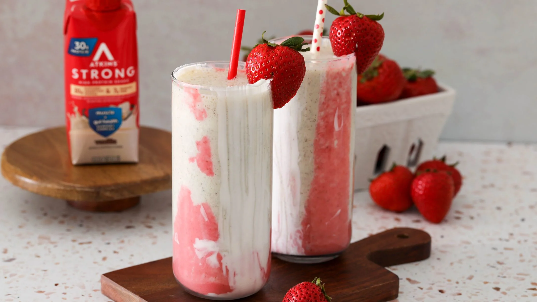Two strawberry smoothies with fresh berries garnish, served on wooden boards with Atkins Strong Shake nearby.