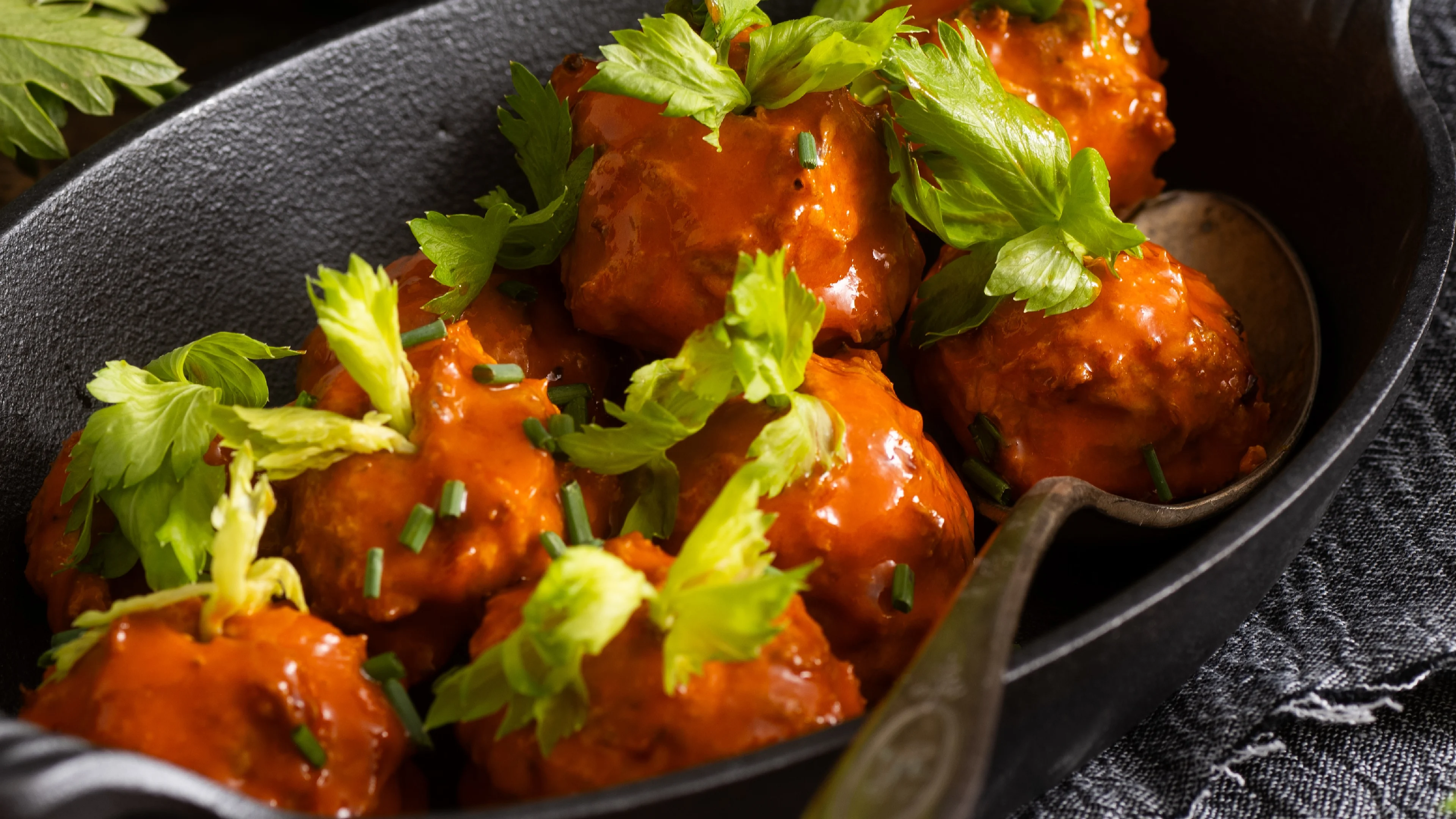 Buffalo meatballs in a cast iron skillet, glazed with bright orange sauce and garnished with fresh celery leaves.