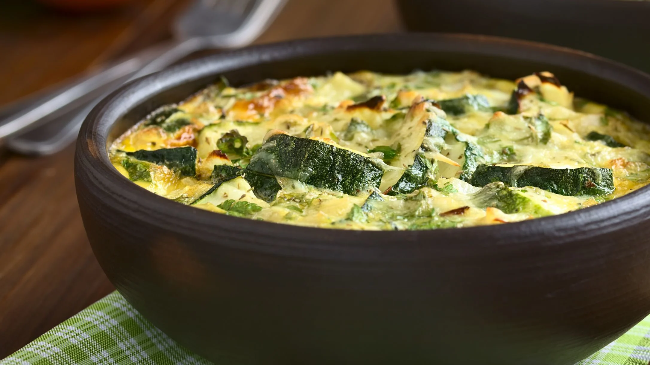 Golden scrambled eggs with zucchini and spinach in a dark ceramic dish, placed on a green checkered cloth.