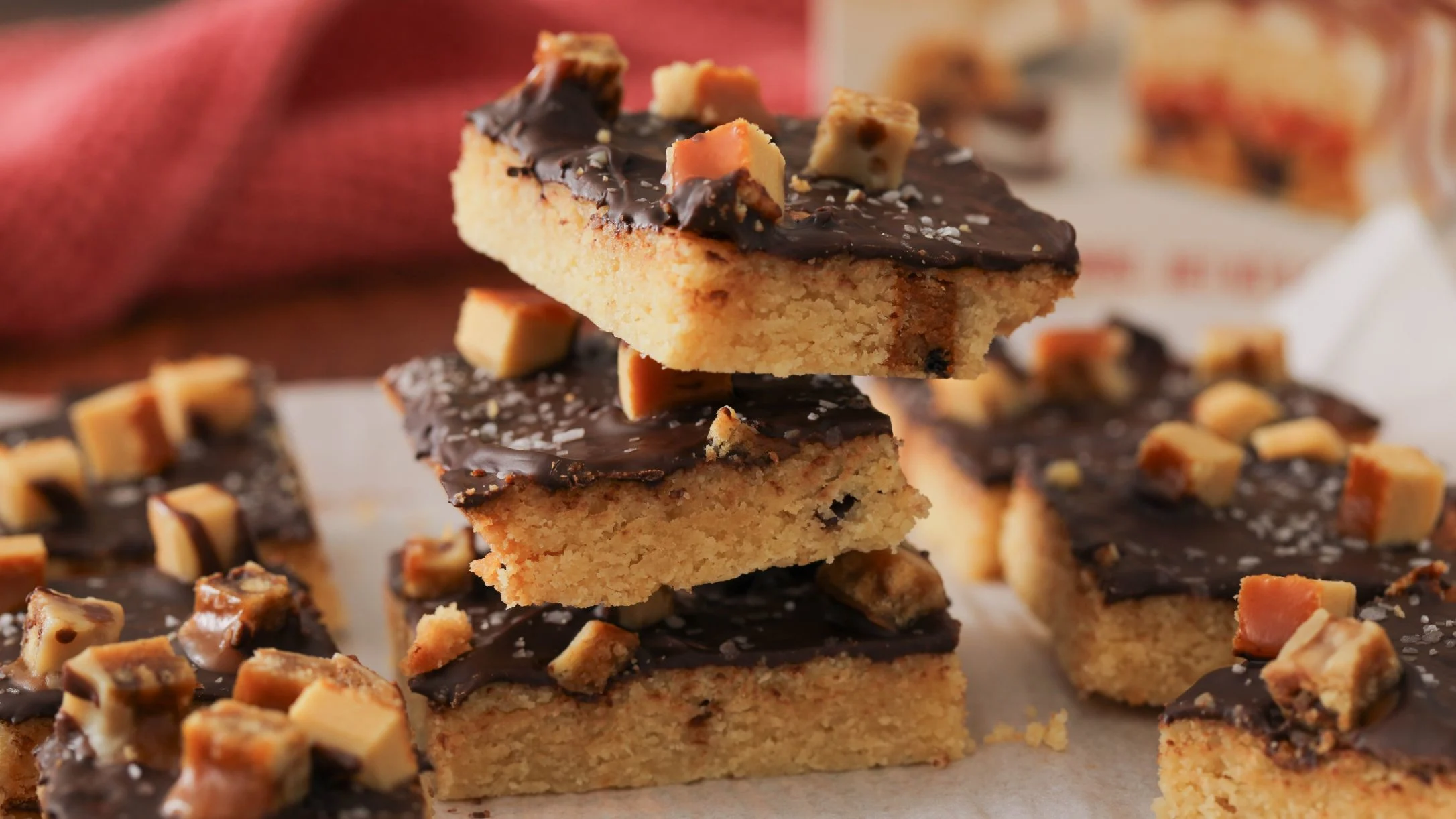 Stack of shortbread cookie bars topped with chocolate ganache and caramel pieces on parchment paper.