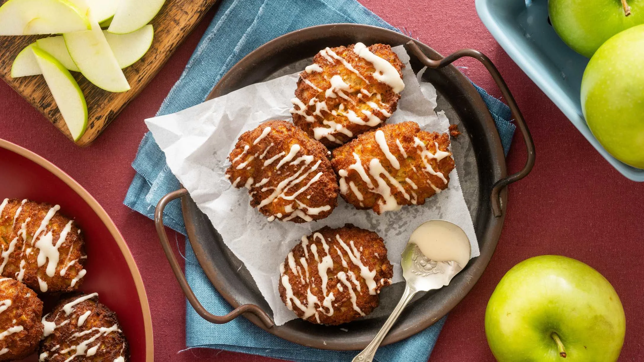 Golden-brown apple fritters drizzled with white glaze on a rustic tray, surrounded by fresh green apples and sliced apple pieces.