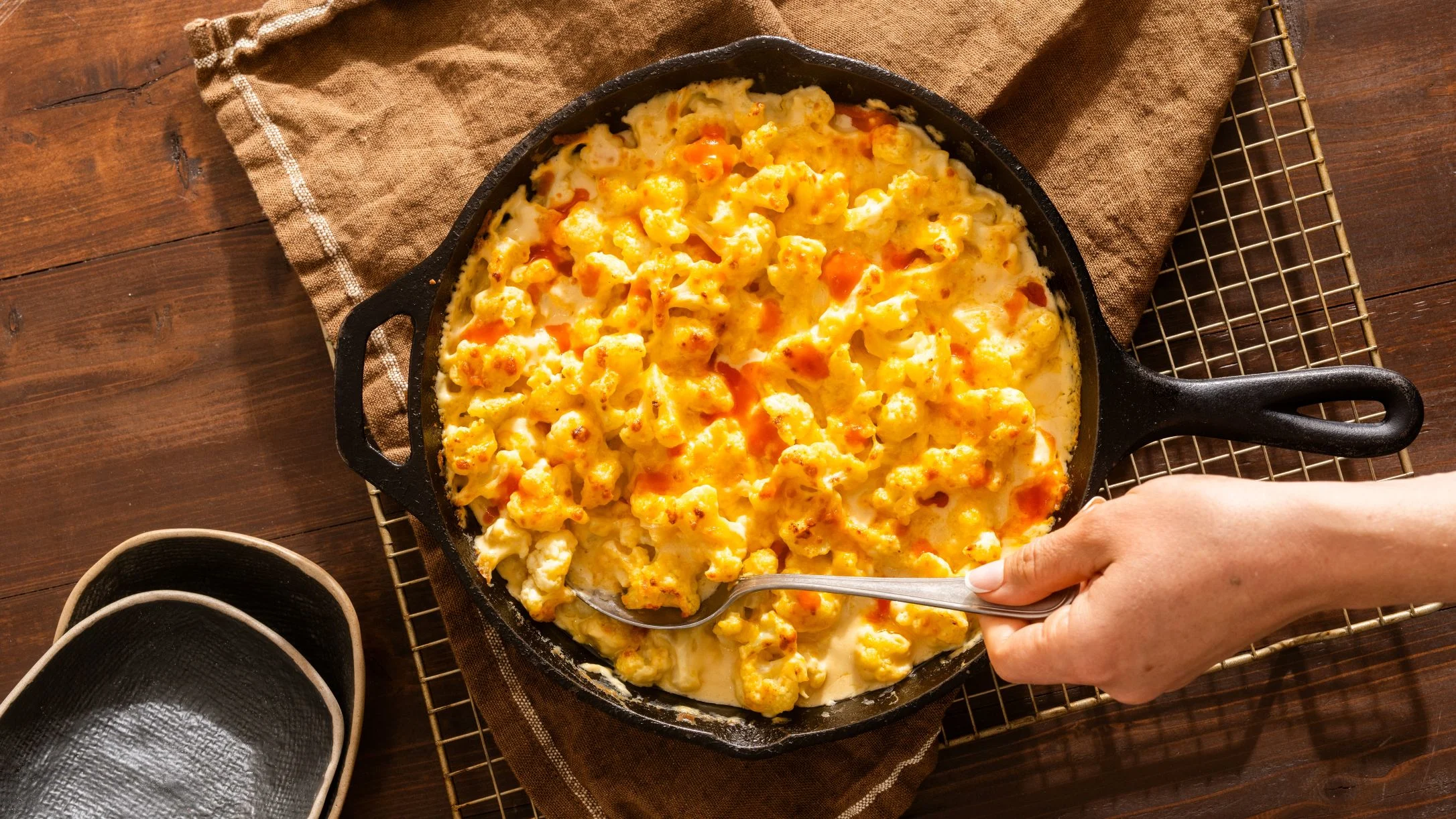 A hand scooping cheesy baked cauliflower from a cast iron skillet on a wooden surface with a tan cloth.