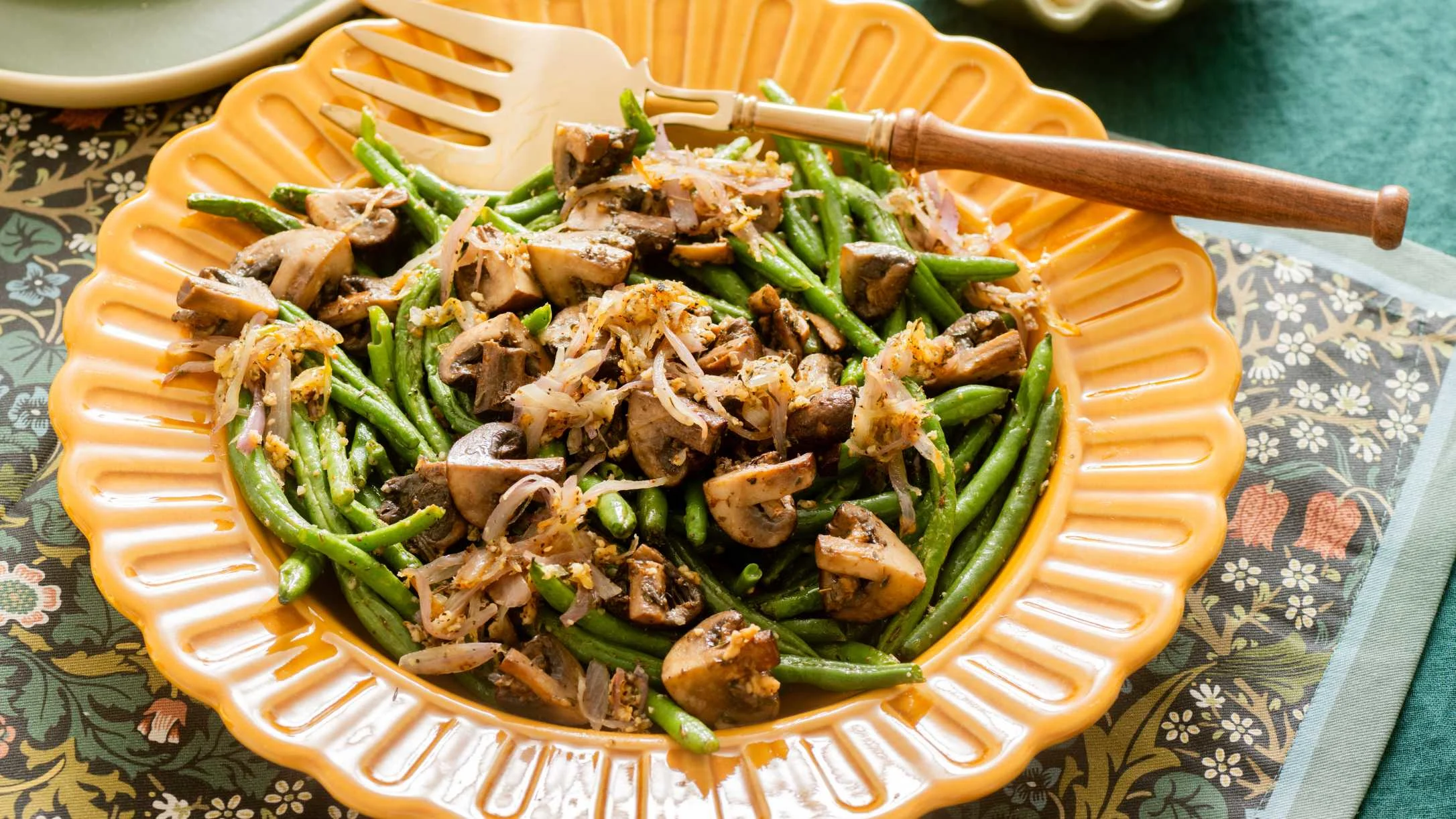 Sautéed green beans and mushrooms with caramelized onions served in a scalloped orange dish with wooden serving utensil.