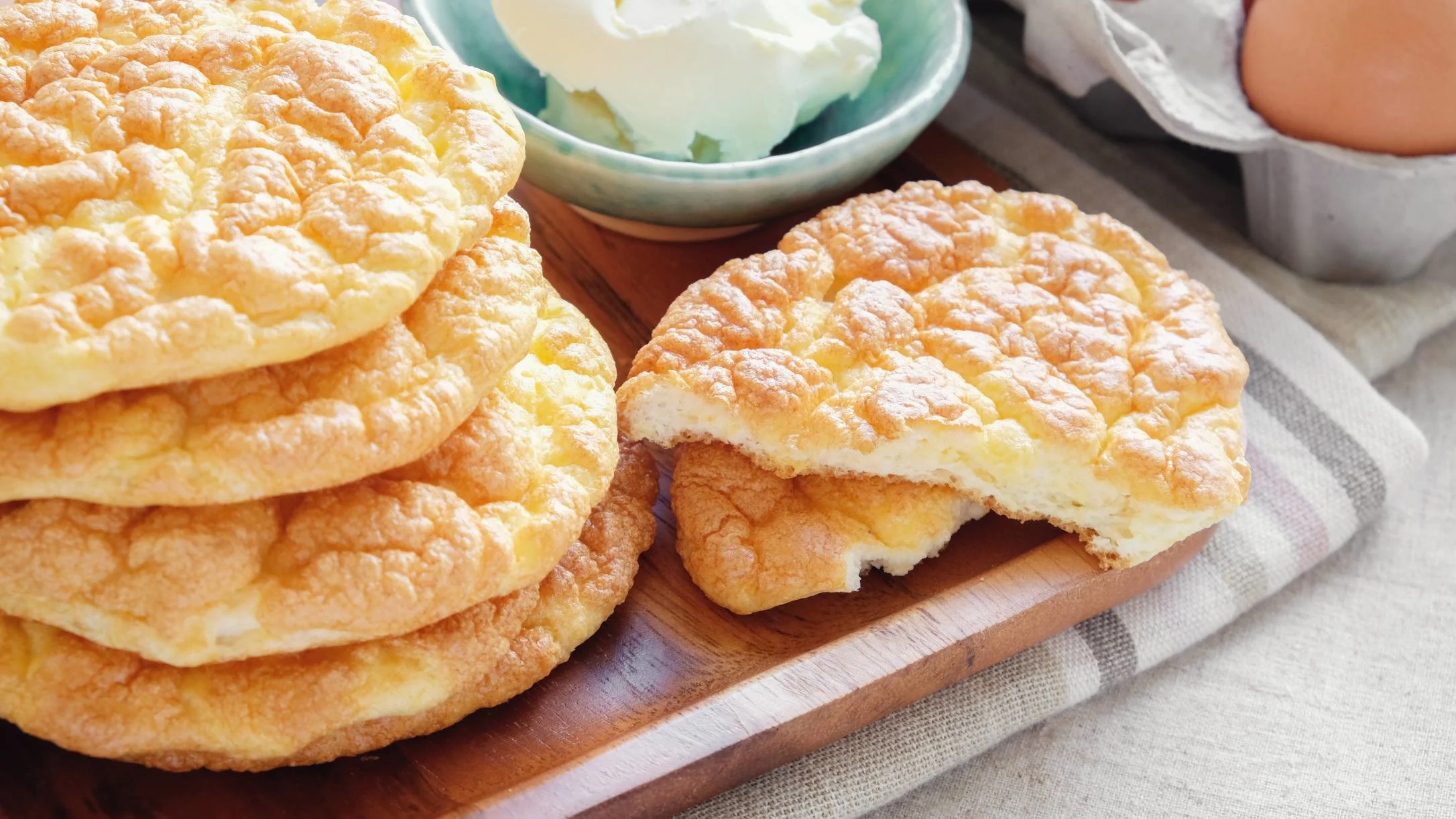 A stack of golden cloud bread on a wooden board, one broken open, with cream cheese in a bowl and eggs nearby.