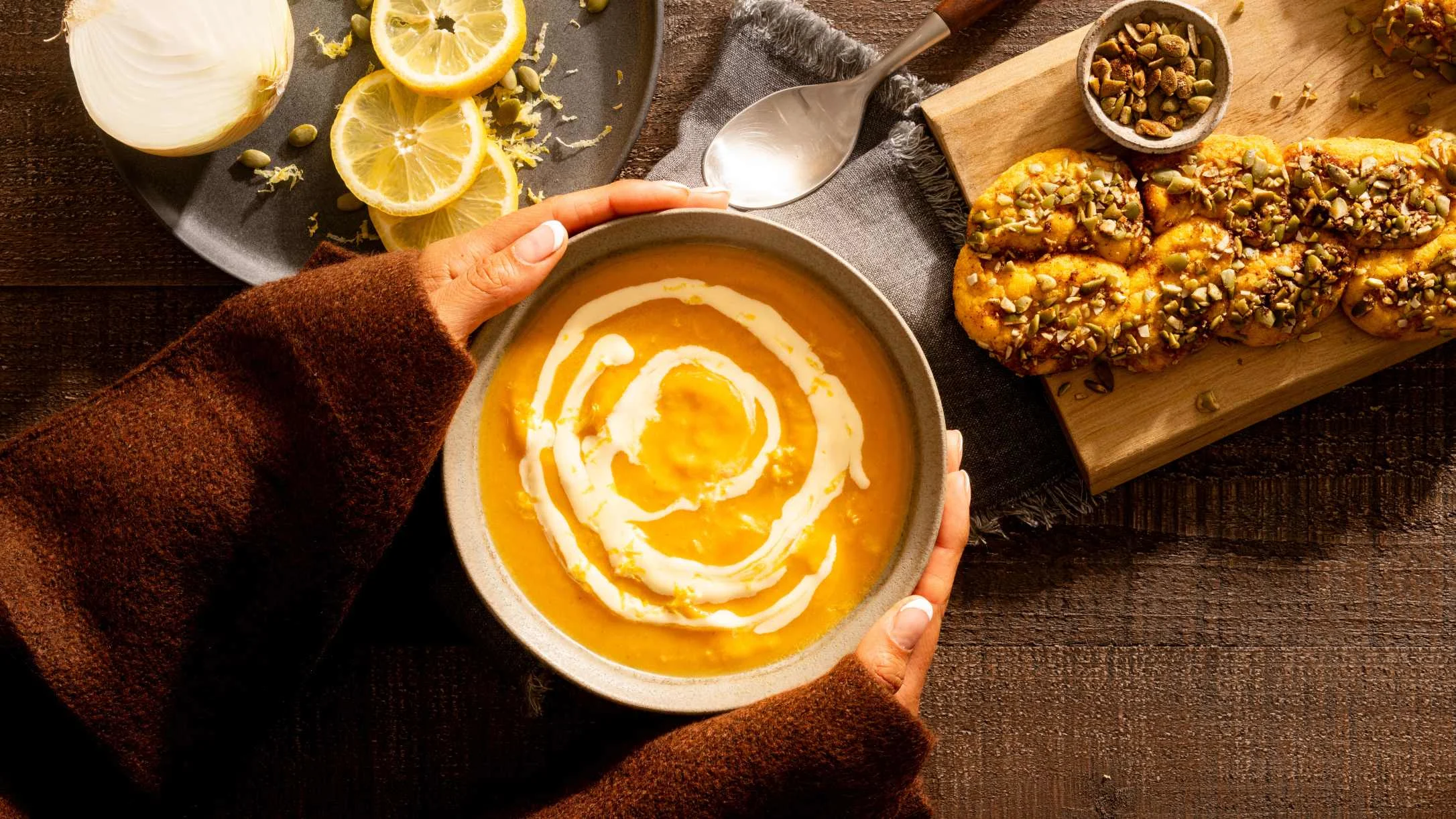Hands holding a bowl of creamy orange soup with a cream swirl, beside seed-topped bread, lemon slices, and an onion.