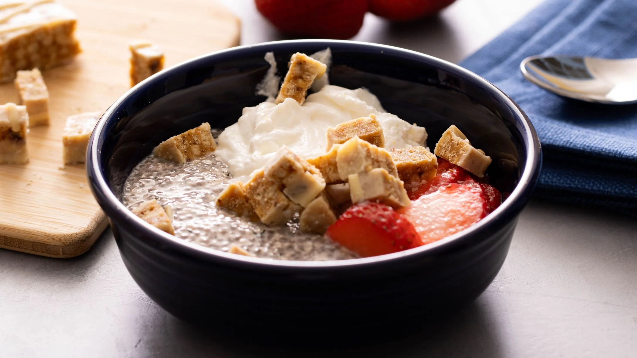 Navy blue bowl containing chia pudding, yogurt, sliced strawberries and protein bar chunks on a wooden board.