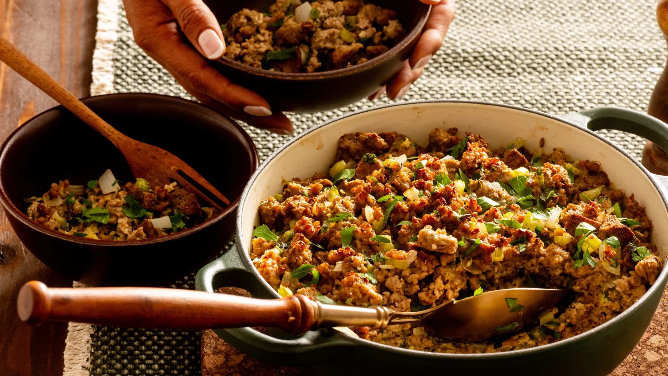 A green casserole dish filled with golden-brown stuffing topped with herbs, alongside serving bowls and wooden utensils.