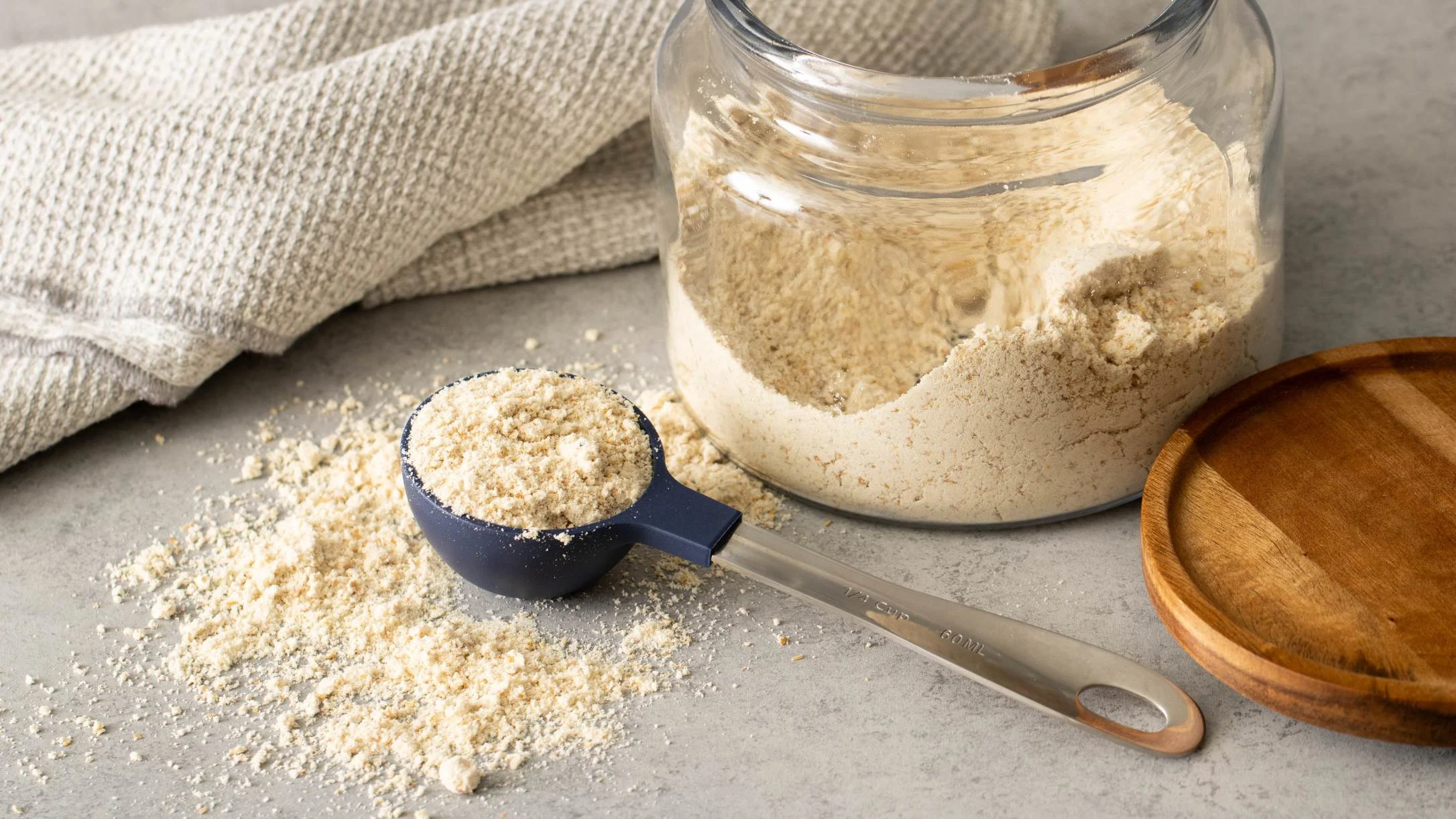 A navy measuring cup filled with flour powder beside a glass jar of flour, a wooden lid, and a linen cloth on a gray surface.