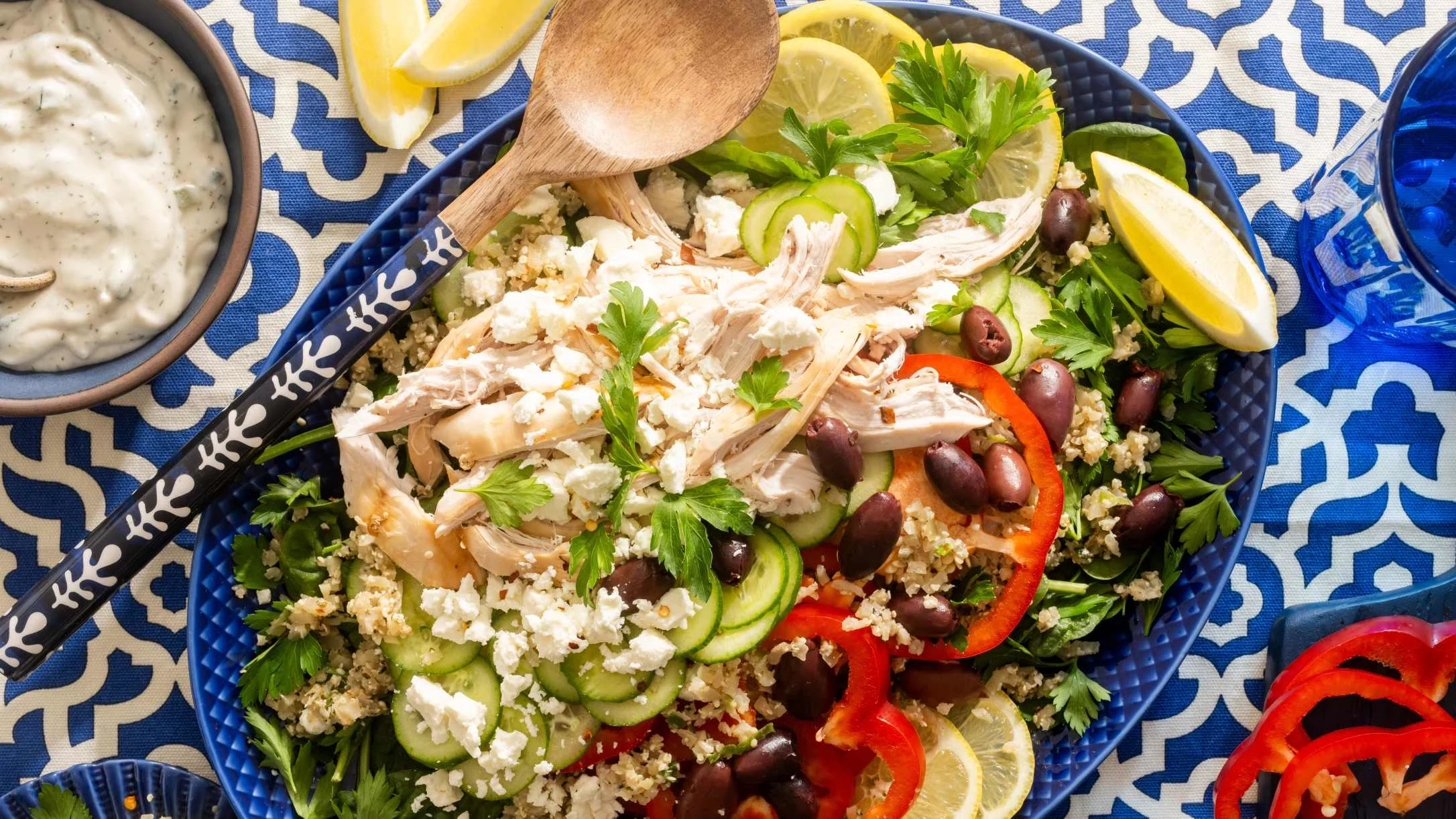 A blue platter with shredded chicken, couscous, feta, olives, cucumber, red pepper, and lemon, with tzatziki on the side.