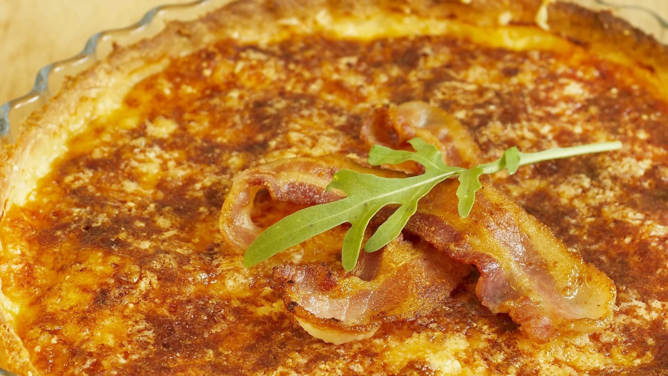 A golden-brown baked tart in a glass dish, topped with crispy bacon strips and fresh arugula leaves as garnish.