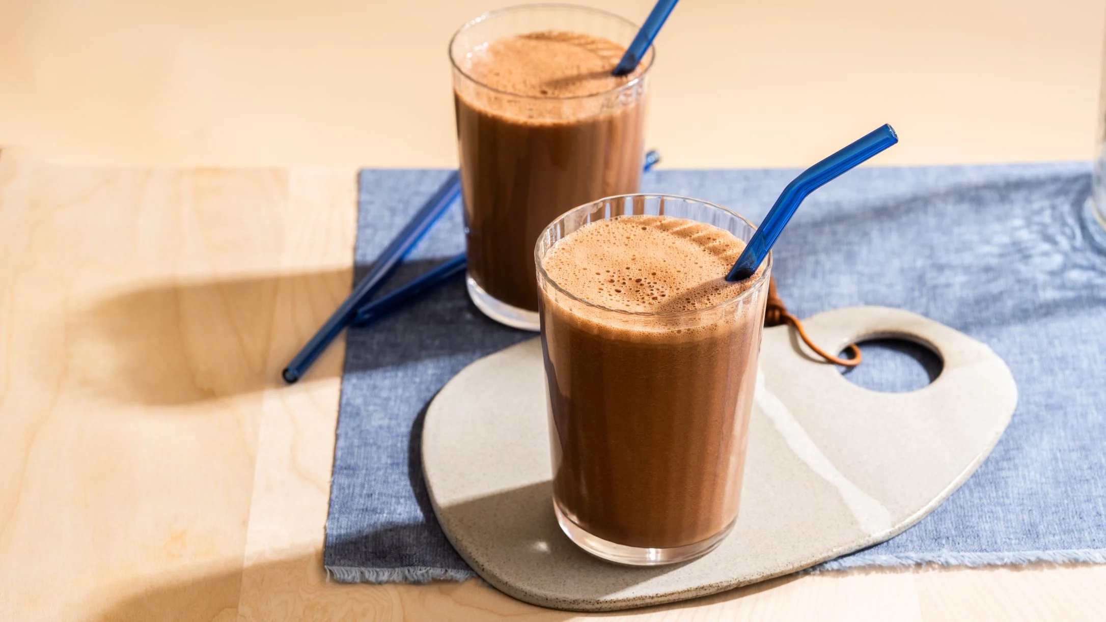 Two tall glasses of chocolate smoothie with blue straws on a ceramic tray with blue cloth on wooden surface.