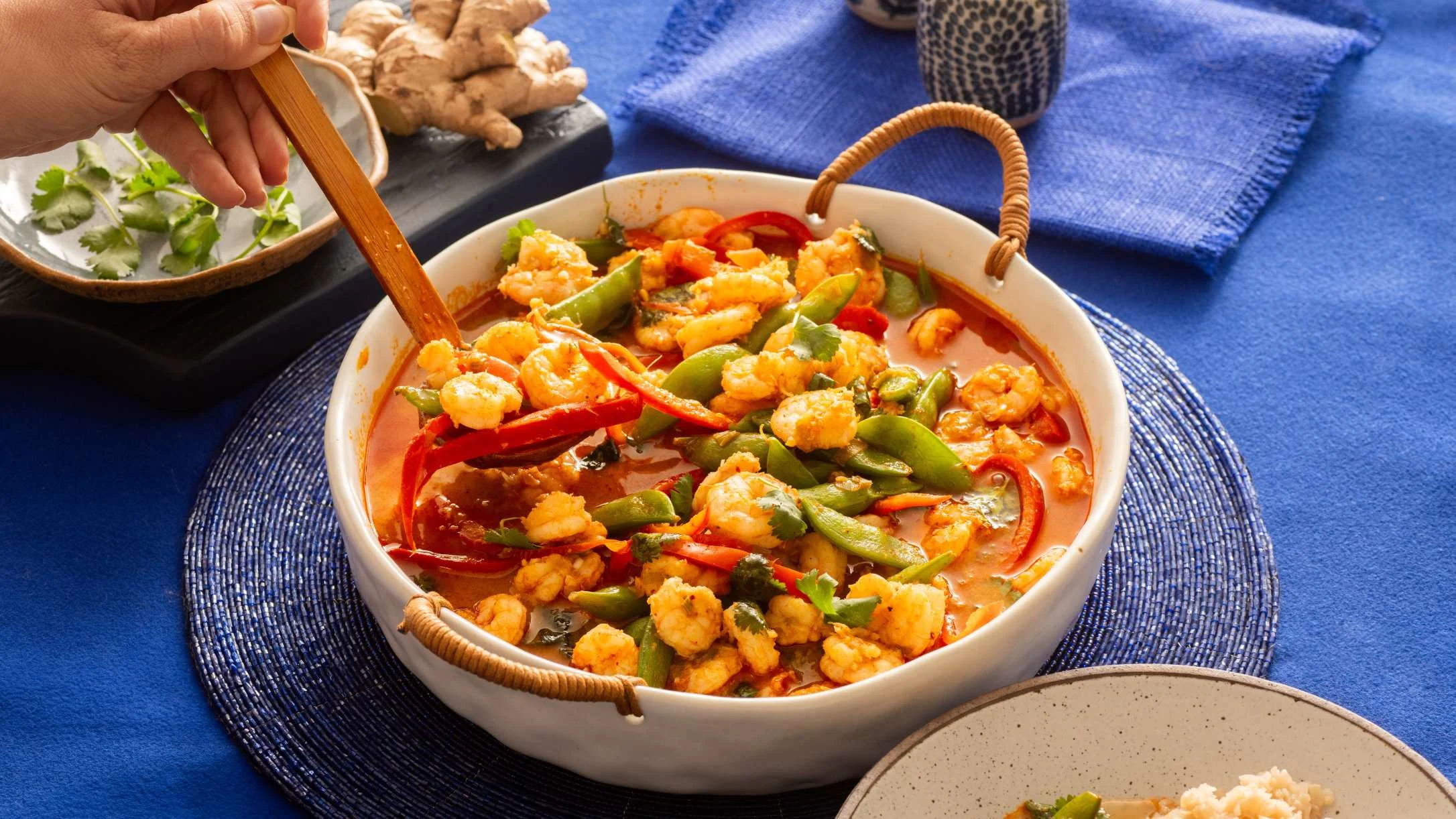 A hand stirs a white bowl of shrimp curry with red and green peppers in a red sauce, served on a blue placemat.