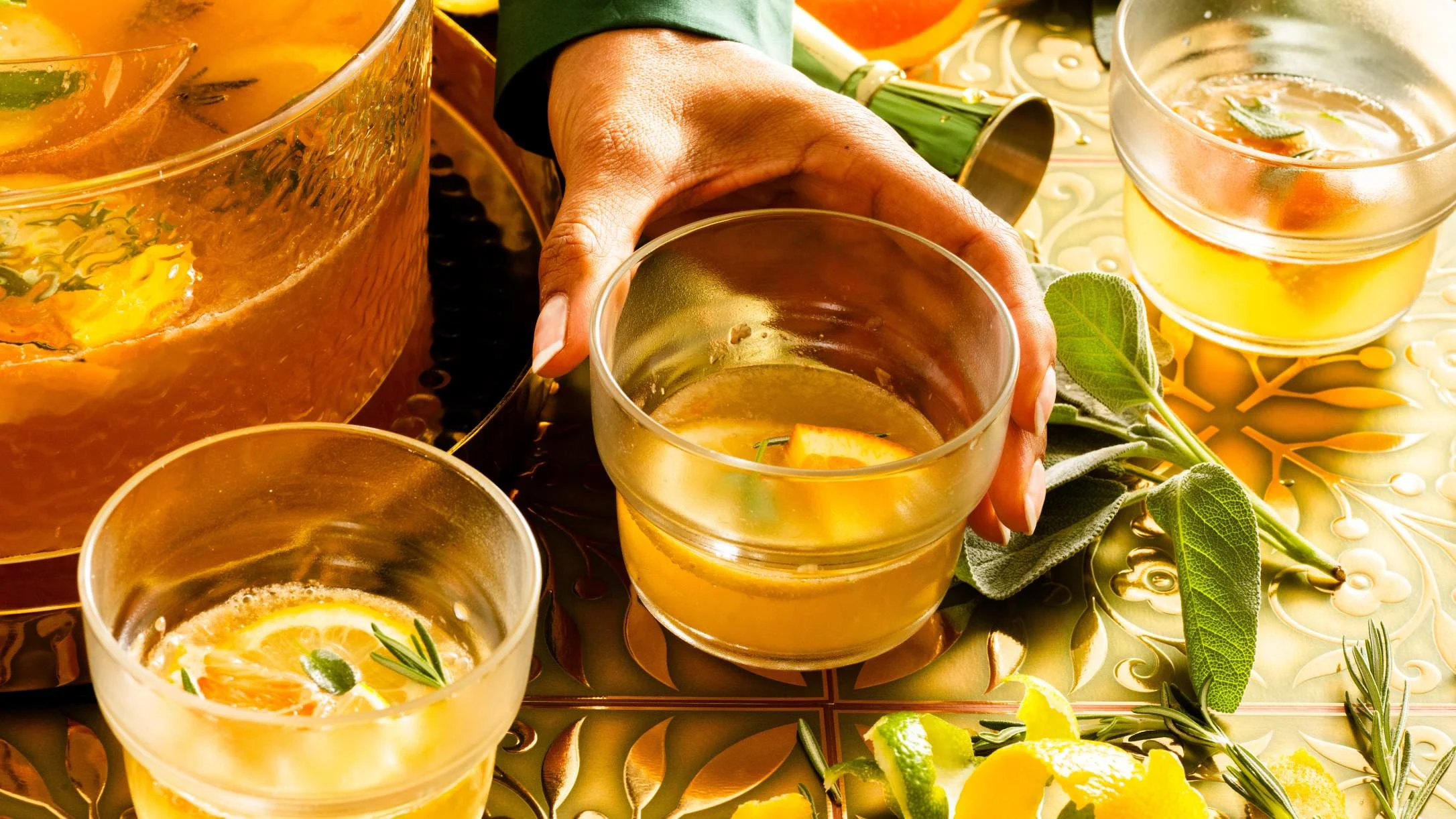 A hand reaching for a glass of golden punch with citrus and herbs, surrounded by more glasses and a large punch bowl.