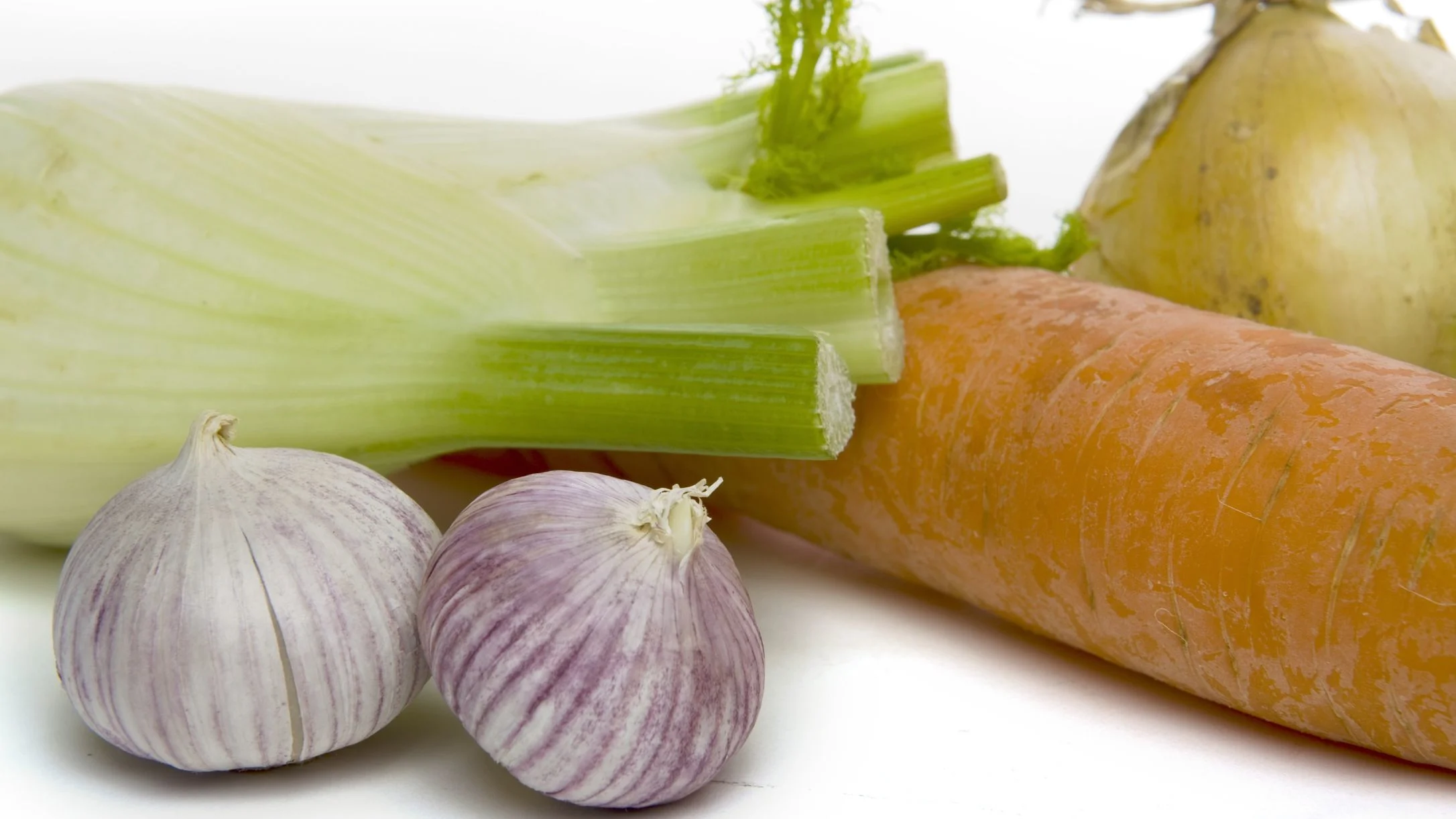 Fresh vegetables on a white surface: fennel bulb, orange carrot, yellow onion, and two purple-striped garlic bulbs.