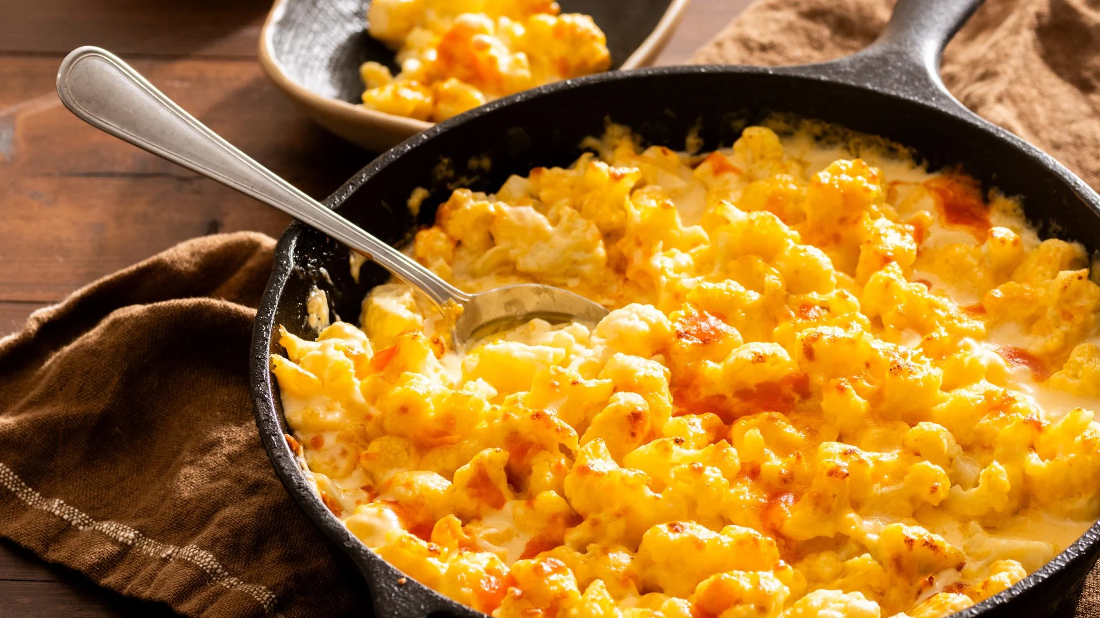 Cheesy baked cauliflower in a cast iron skillet with a spoon, golden and bubbly, served on a wooden surface.