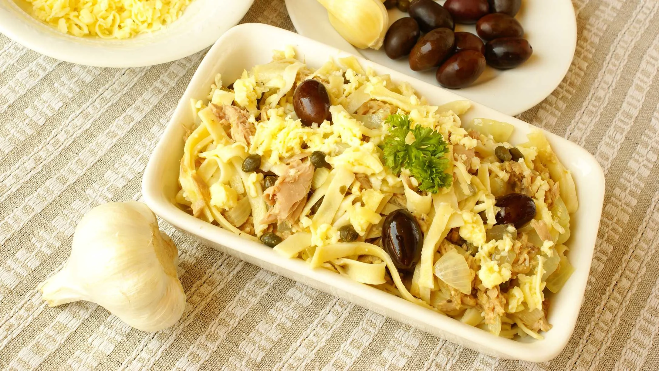 A white dish of pasta with tuna, black olives, capers, and grated egg, garnished with parsley, alongside garlic and olives.