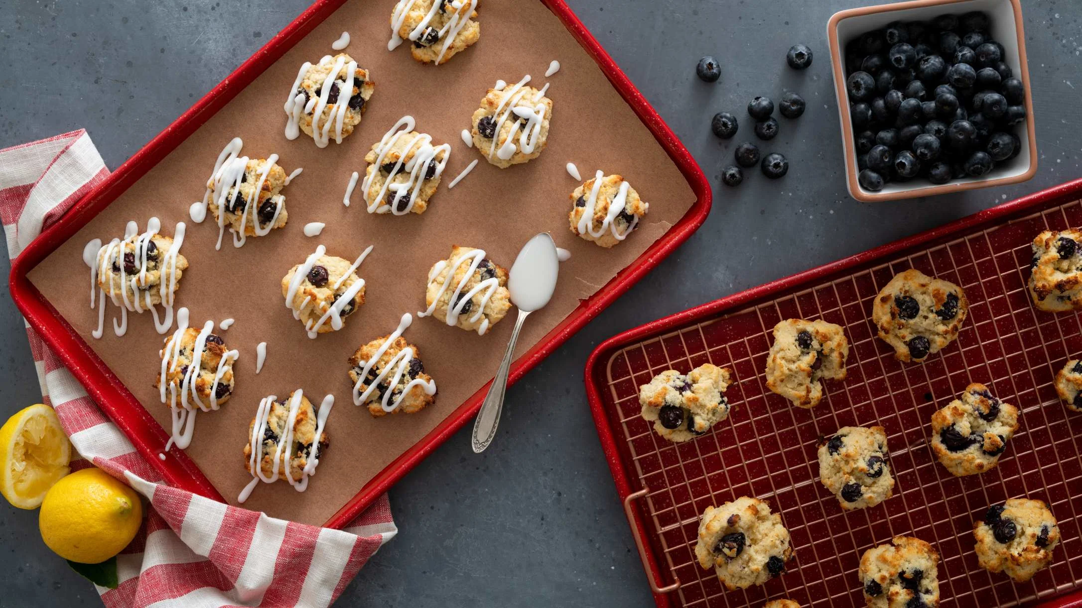 Blueberry scones on red baking trays - some with white icing drizzle, fresh blueberries and lemons nearby.