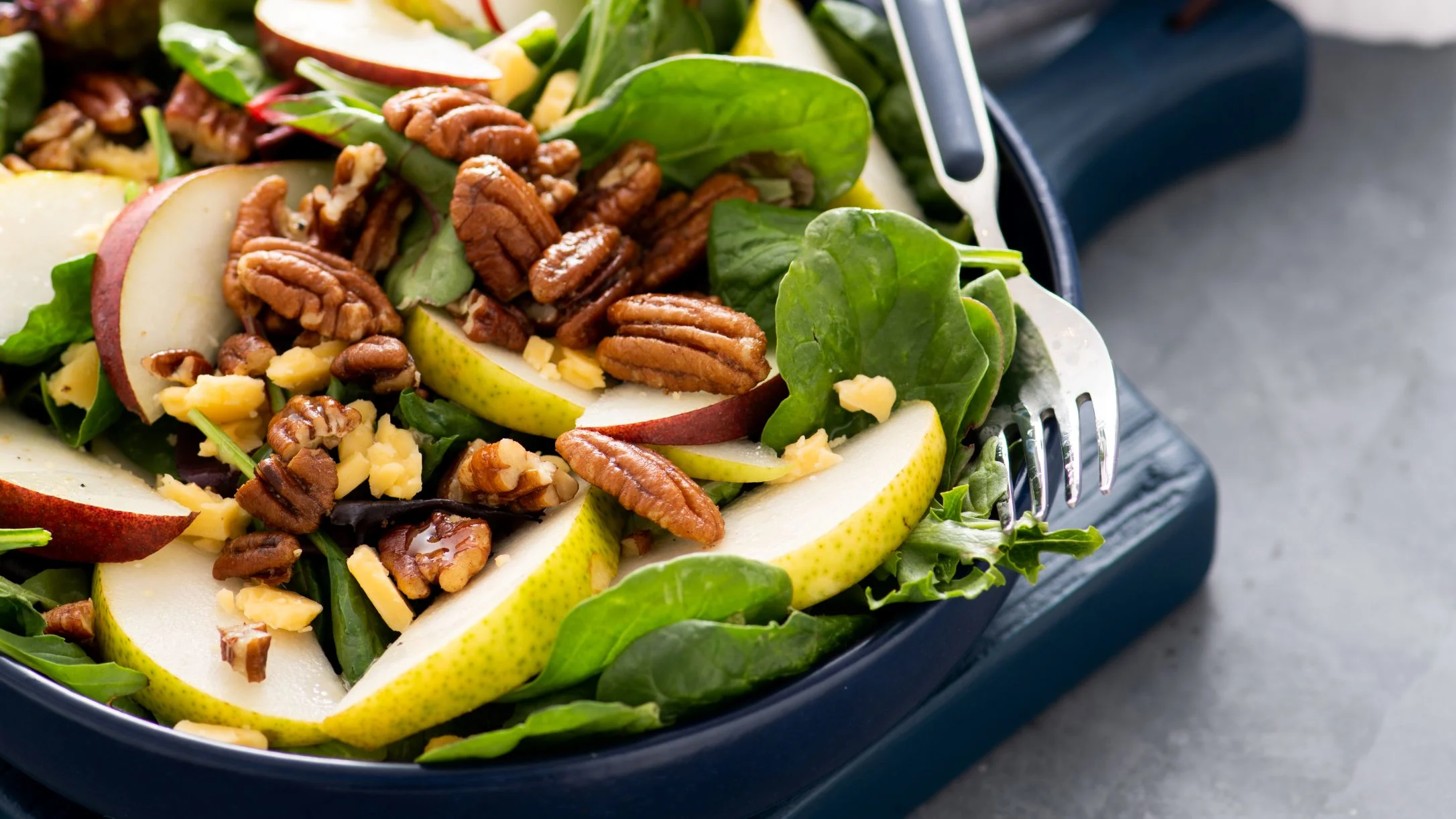 Fresh salad with sliced pears, pecans, cheese, and spinach leaves in a dark blue bowl with a silver fork.