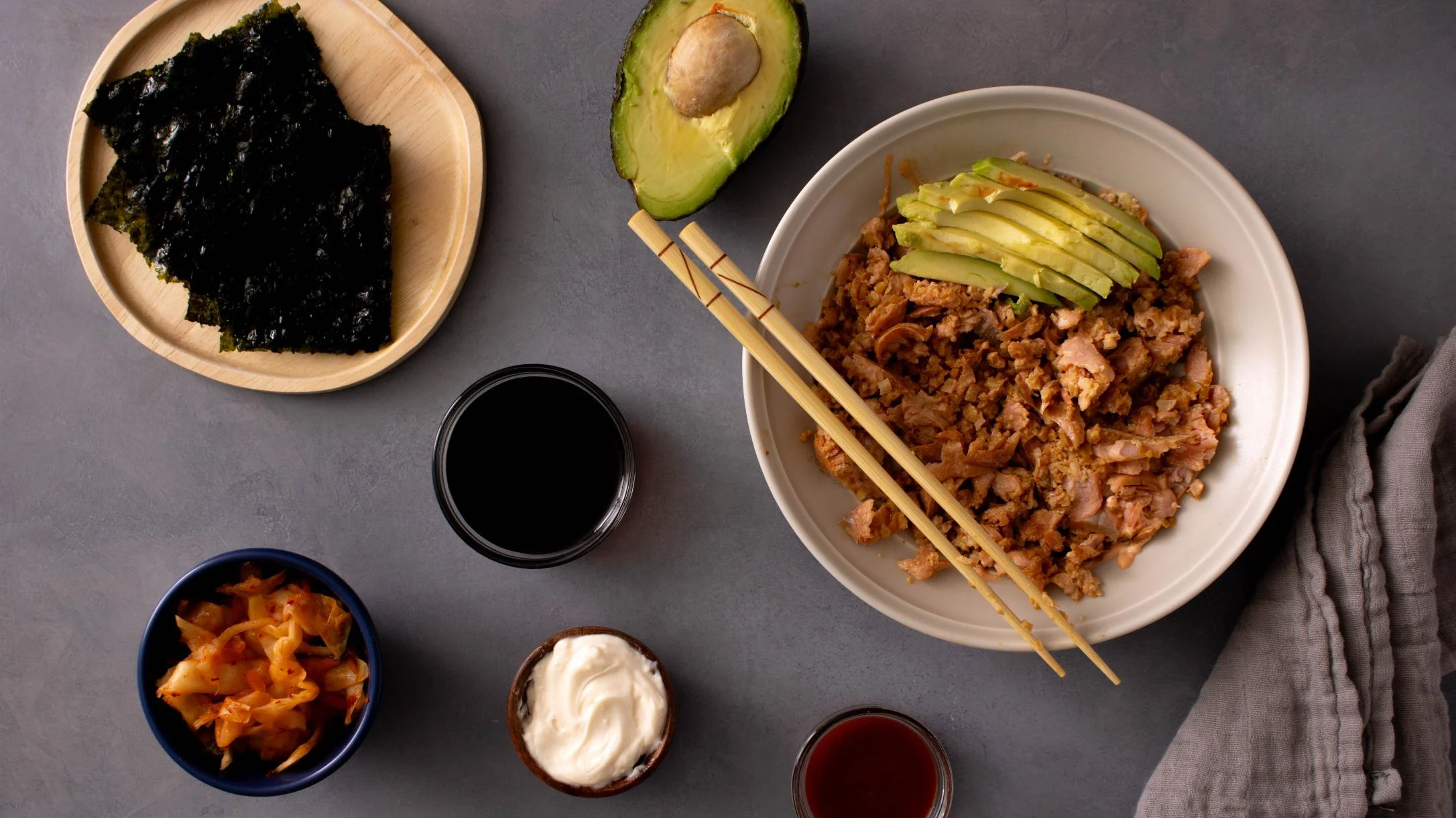 A bowl of salmon and cauliflower rice topped with avocado slices and chopsticks, surrounded by nori, kimchi, soy sauce, and mayo.