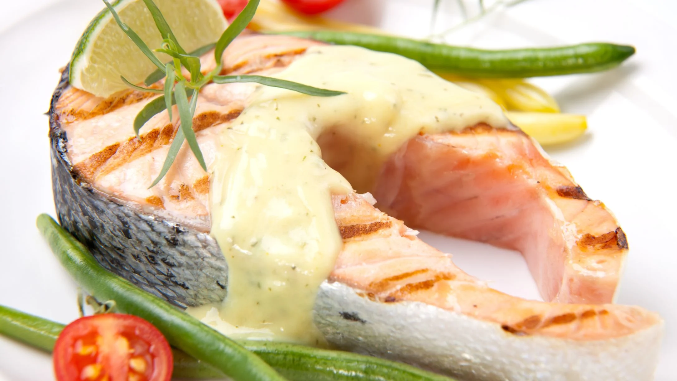 Grilled salmon steak topped with creamy sauce, served with green beans, cherry tomatoes, and a lime wedge on a white plate.
