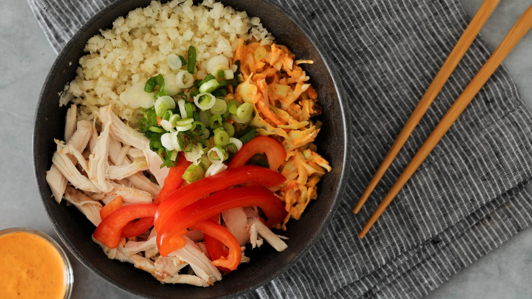 Asian-style rice bowl with shredded chicken, red bell peppers, green onions, and orange sauce, served with chopsticks.