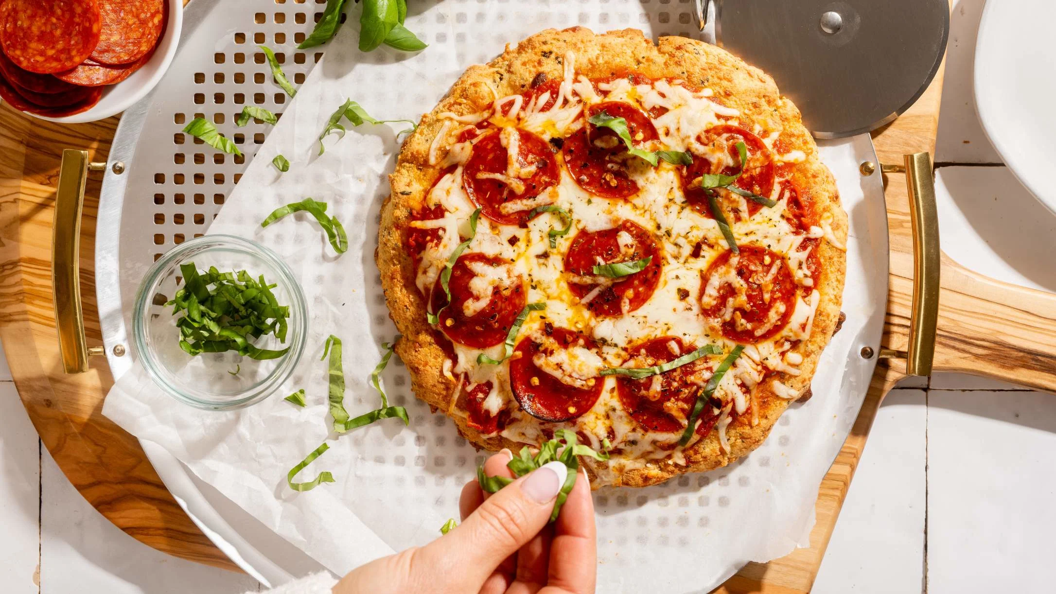 Homemade pepperoni pizza on a perforated tray with melted cheese and fresh basil, hand sprinkling herbs on top.