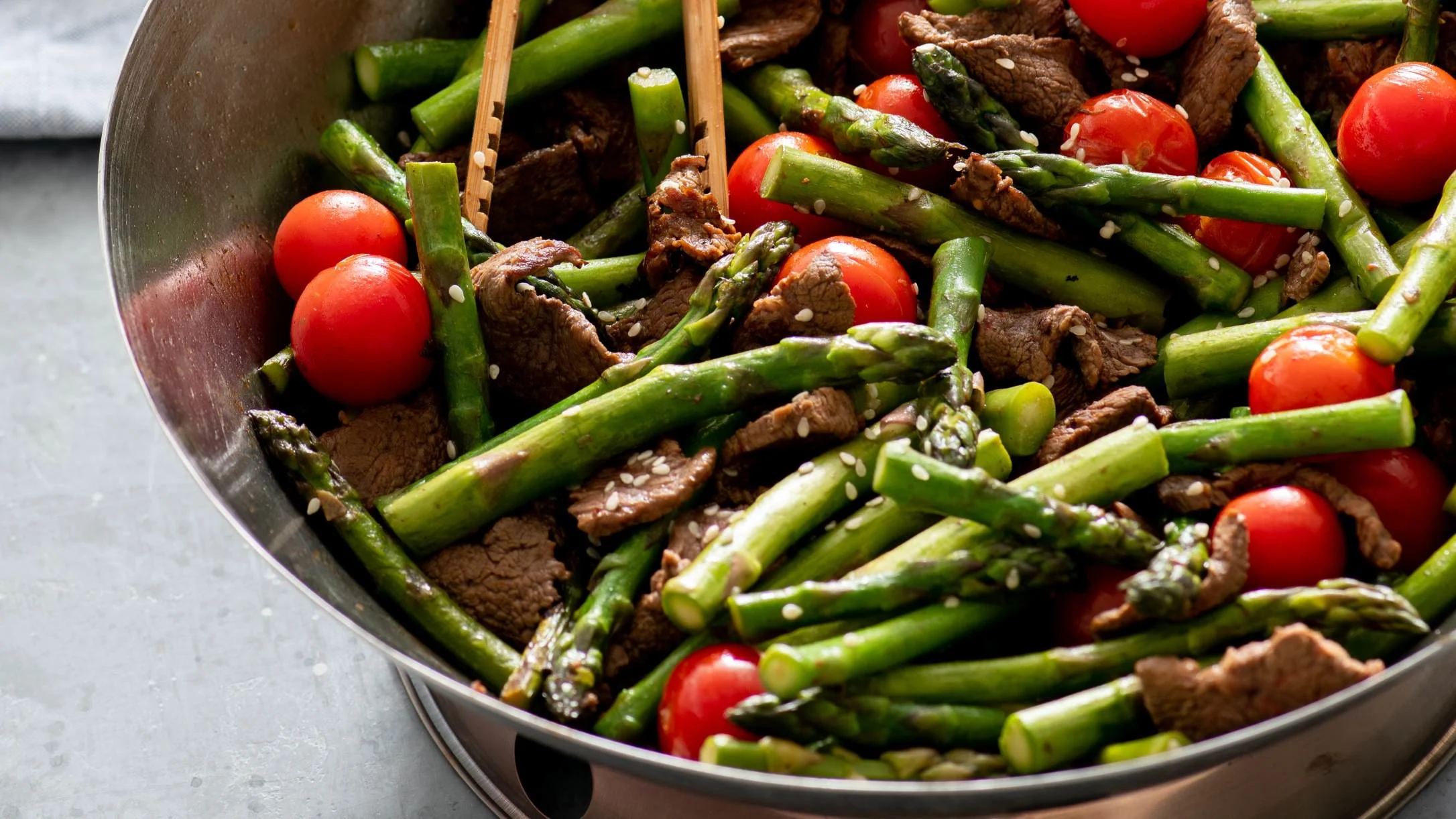 A stir-fry of beef strips, green asparagus, and cherry tomatoes topped with sesame seeds in a steel wok with wooden tongs.