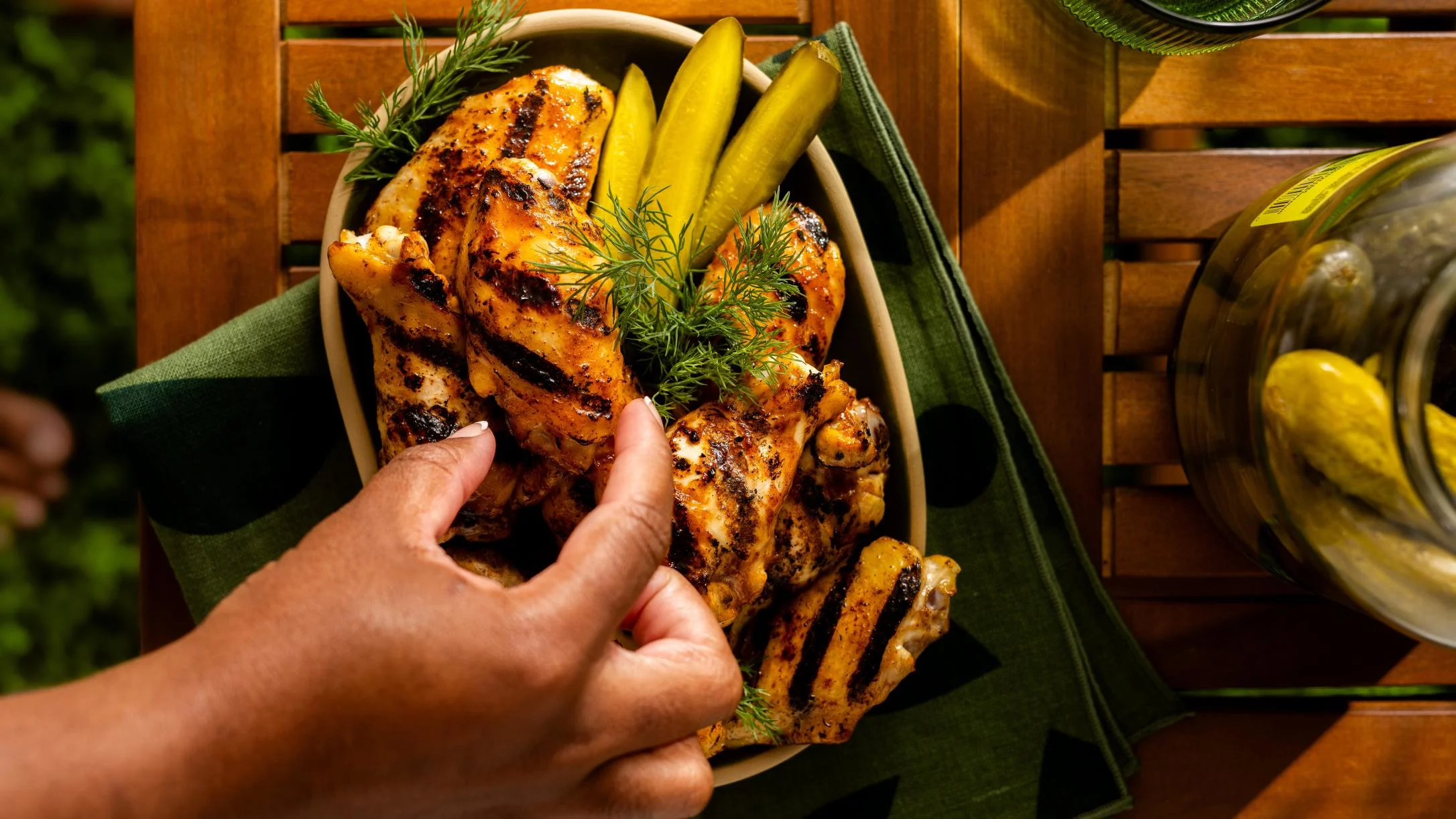A hand reaching for grilled chicken pieces in a bowl garnished with dill and pickles, placed on a green napkin on a wooden surface.