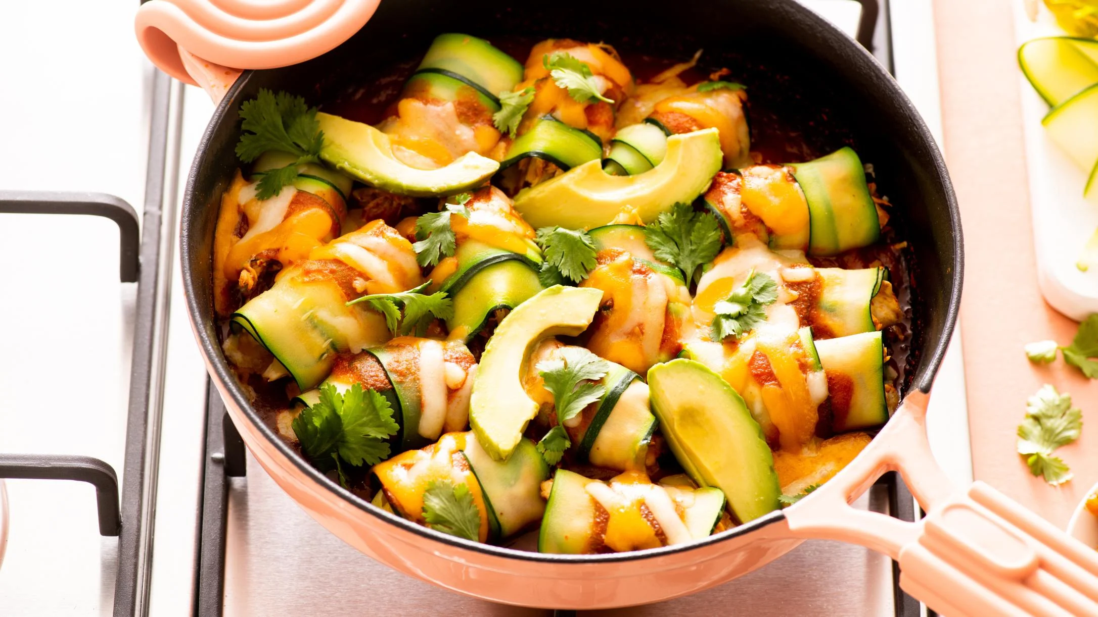 Copper pan filled with zucchini-wrapped enchiladas topped with melted cheese, avocado slices, and fresh cilantro.