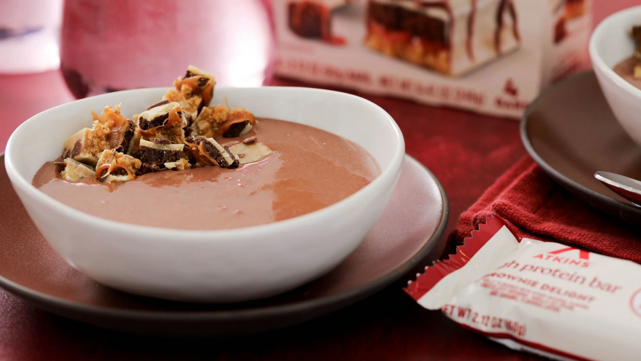 Creamy chocolate pudding in white bowl topped with candy bar pieces, served on pink plate with protein bar wrapper nearby.
