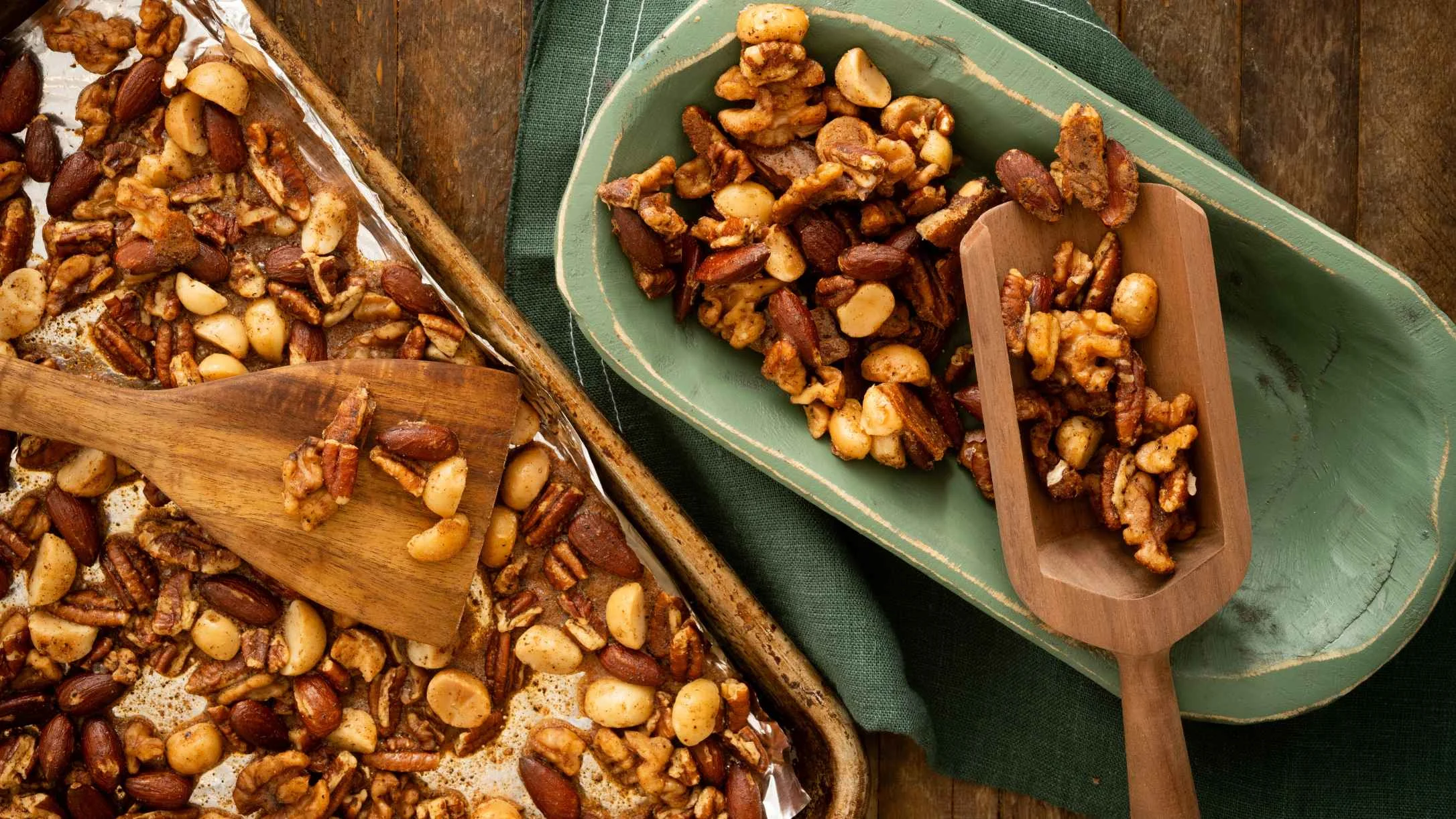 Roasted mixed nuts with almonds, pecans, and macadamias on a baking sheet and green wooden tray with a wooden scoop.