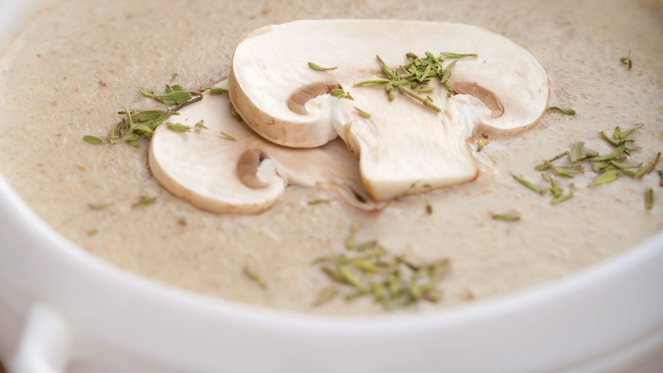 Creamy mushroom soup in a white bowl, topped with sliced mushrooms and scattered fresh green herbs.