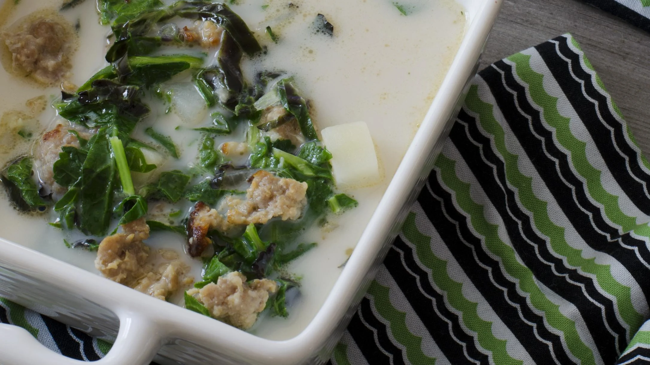 A white bowl of creamy soup with kale, sausage, and rutabaga slices, beside a green and black striped napkin.