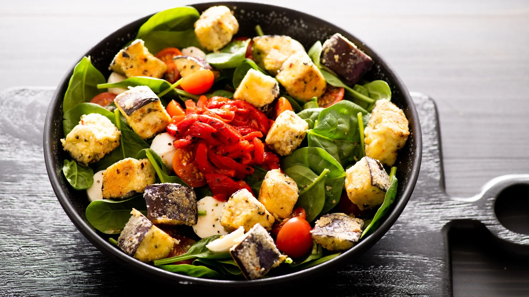 Fresh salad in black bowl with spinach, eggplant, cherry tomatoes, and roasted red peppers on wooden table.