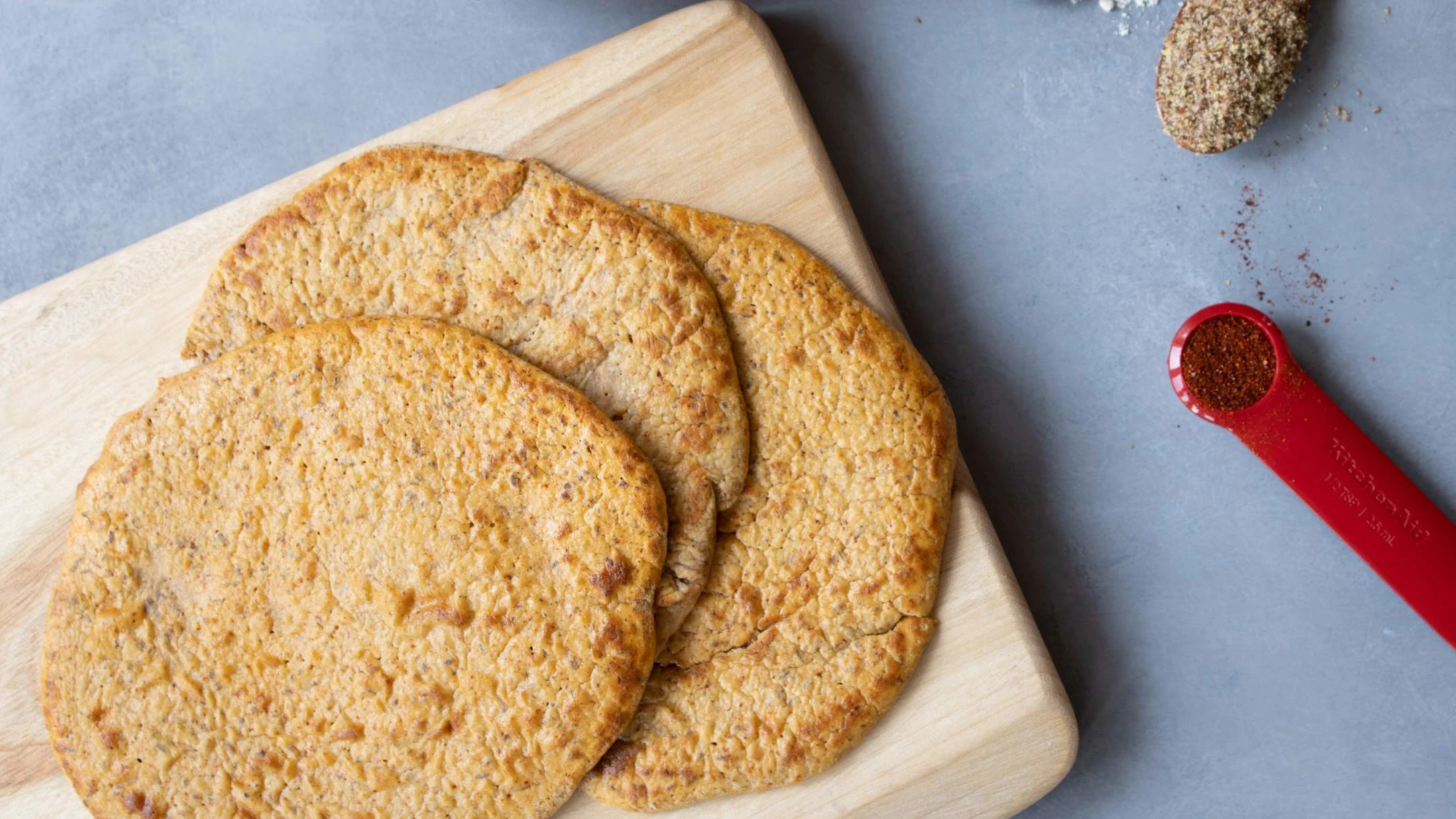 Three golden-brown flatbreads stacked on a wooden cutting board, with a red measuring spoon of spice nearby.